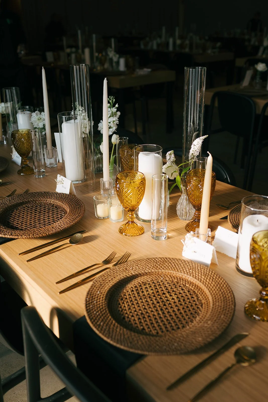 Elegant dining table set with woven placemats, amber glasses, white candles in various holders, white flowers, and silverware, preparing for a formal event.