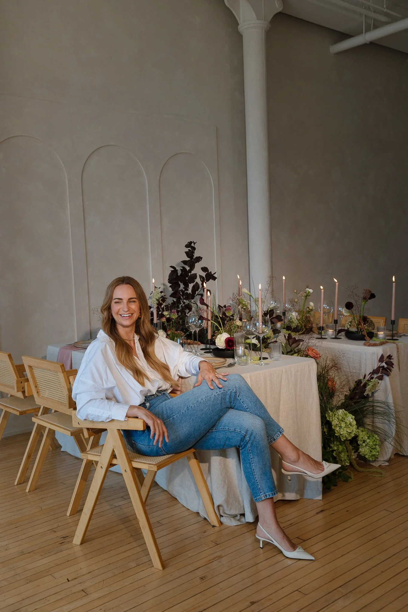 A woman with long wavy hair sitting on a wooden chair, smiling, in front of a decorated dining table with candles and flowers, in an elegant room with textured walls and a wooden floor.