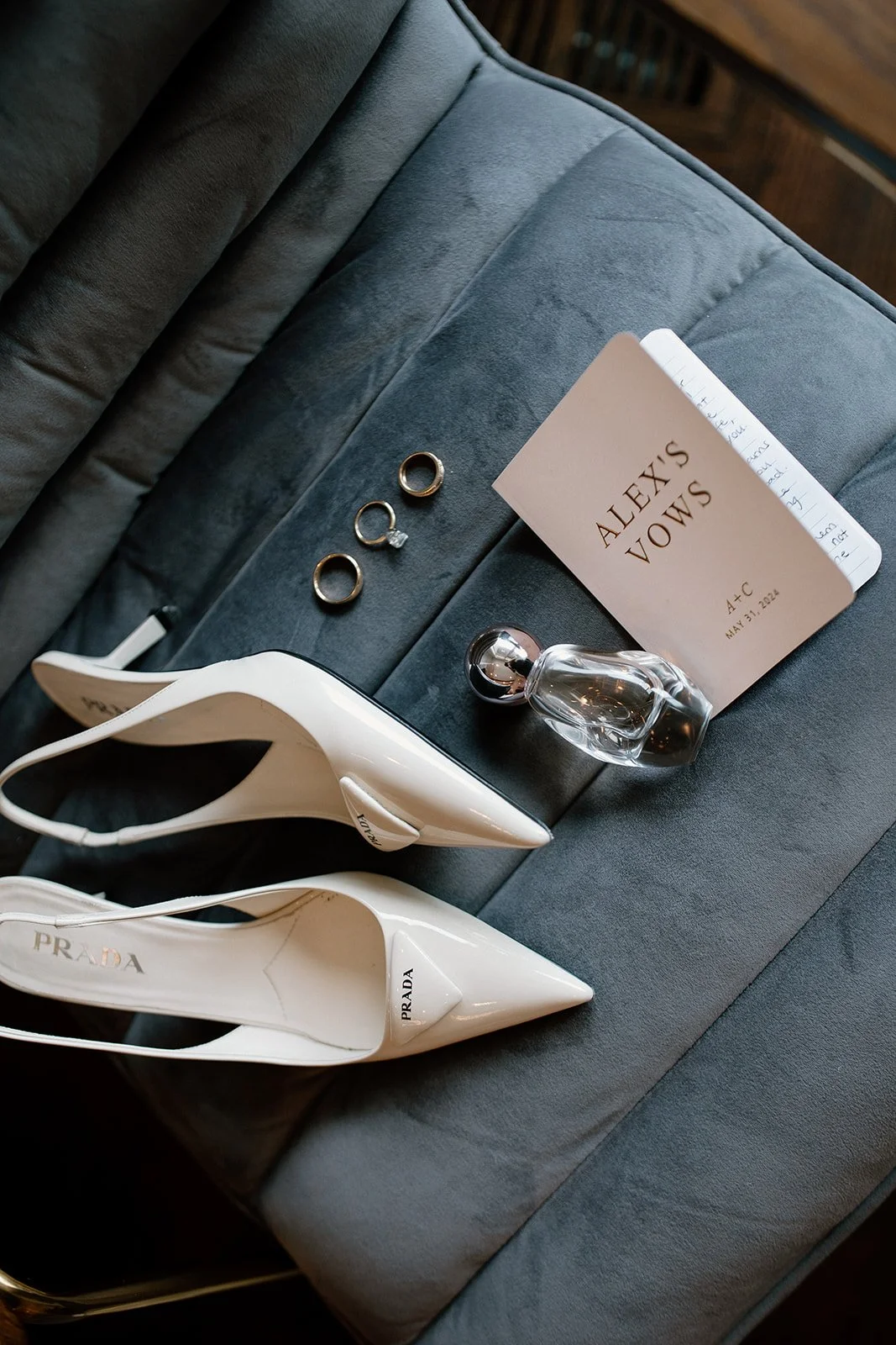 A pair of white Prada high heels, three rings, a perfume bottle, a card labeled 'Alex's Vows', and a notebook on a grey upholstered surface.