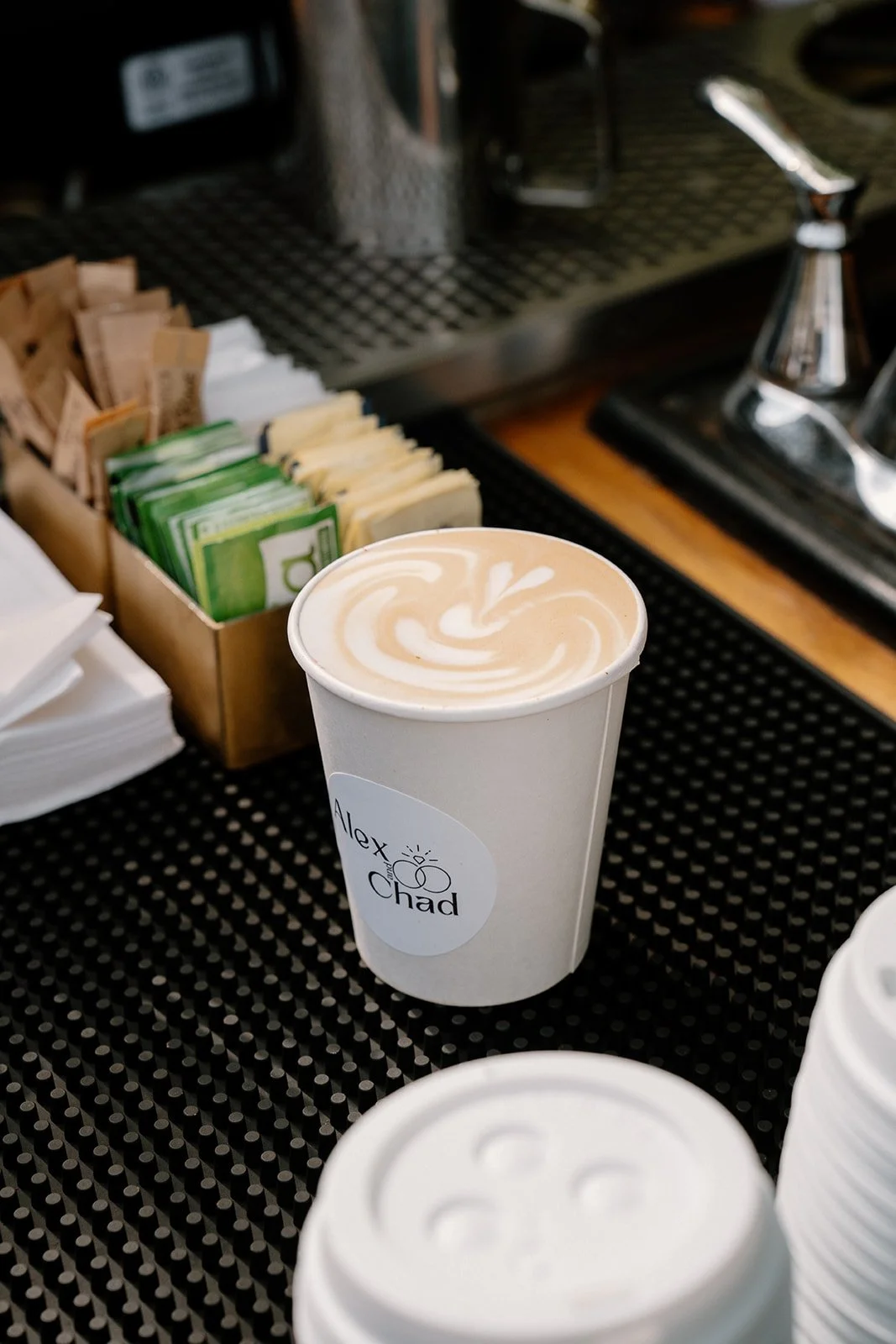 A paper cup of coffee with latte art resting on a black textured surface, with coffee stirrers, sugar packets, and a coffee machine in the background.