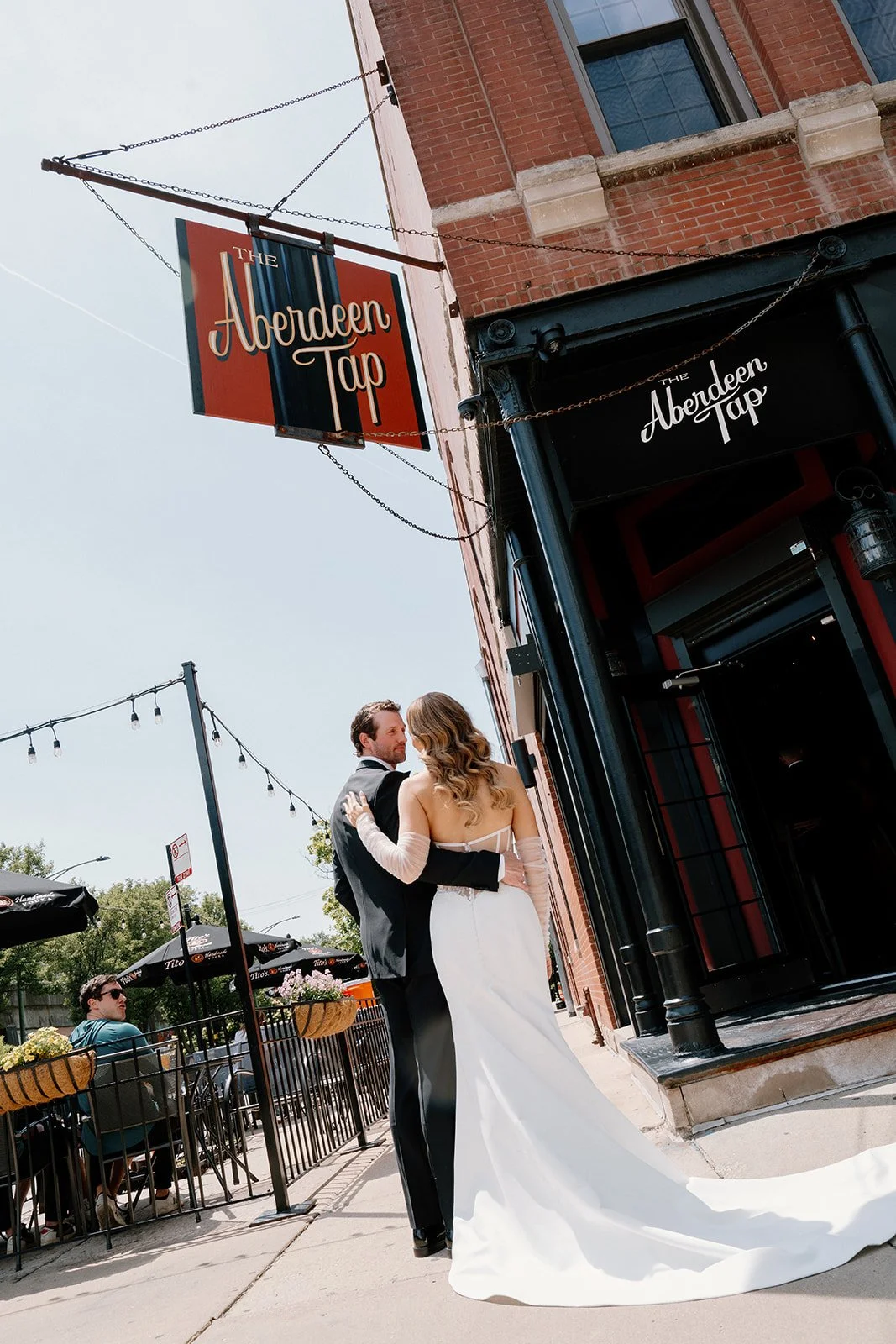 A couple dressed in wedding attire dancing outside The Aberdeen Tap bar, with outdoor seating and string lights overhead.