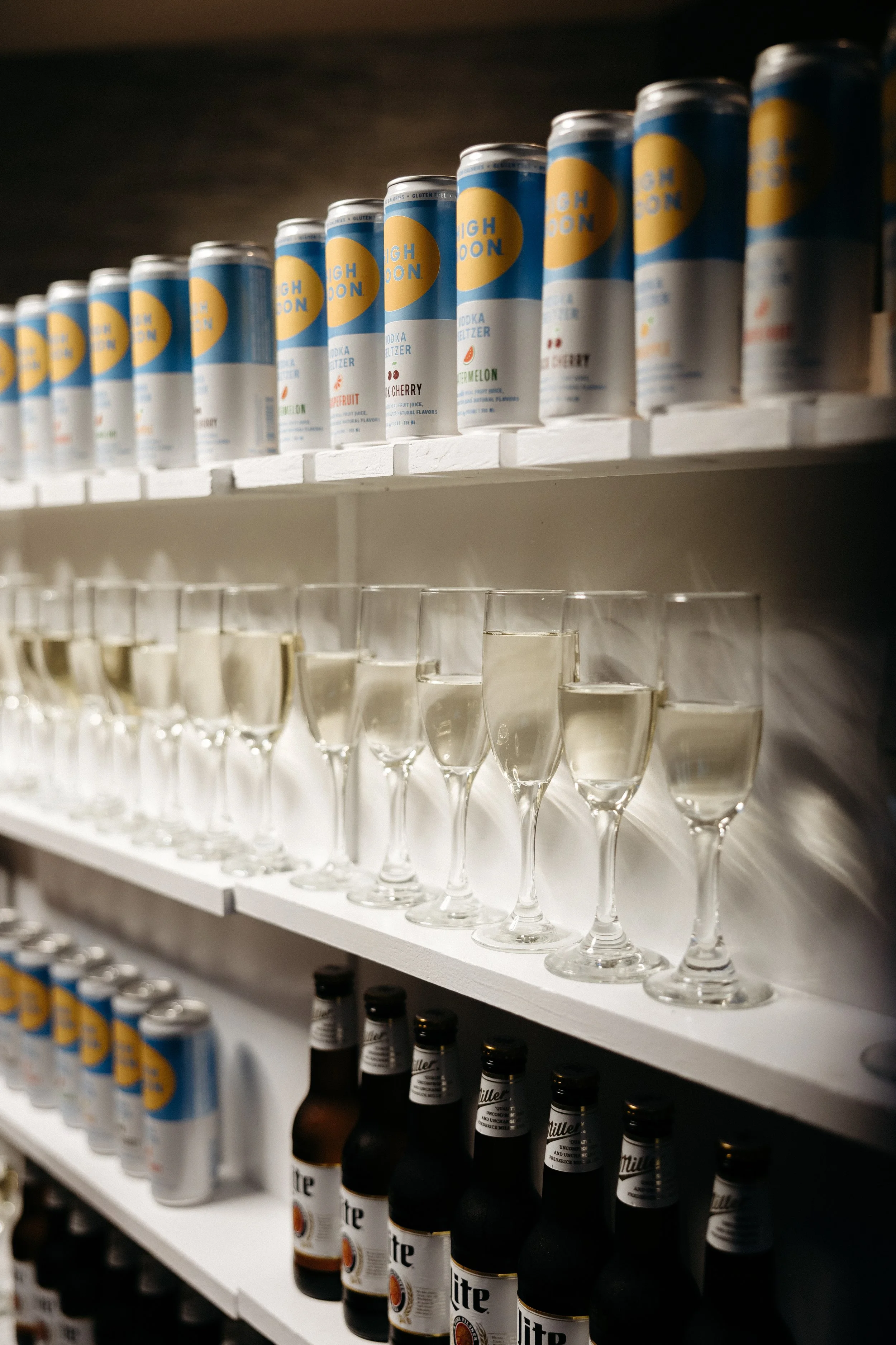 Shelves with canned High Noon vodka seltzers, glass of champagne, and Miller Lite beer bottles.