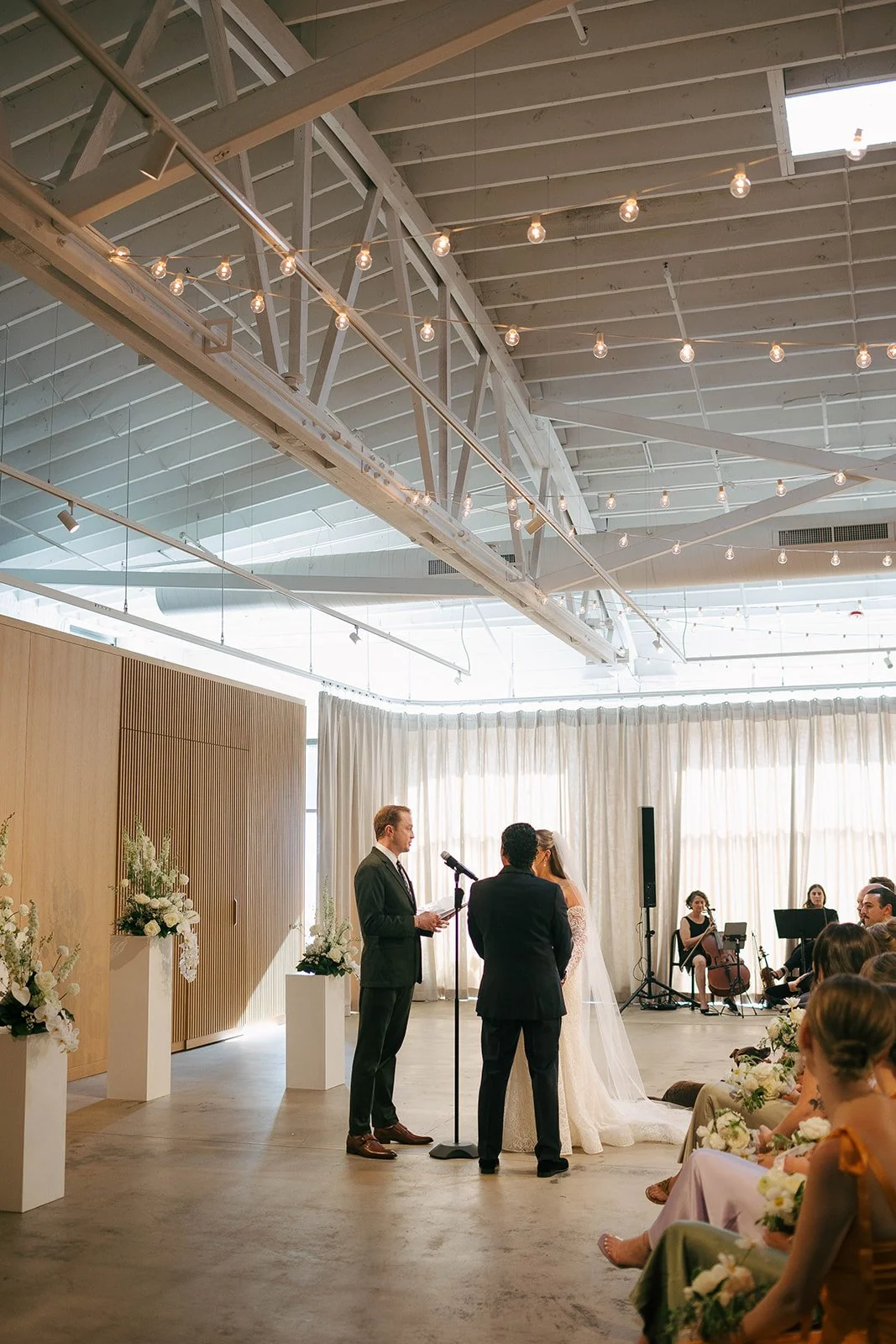 A wedding ceremony in a modern, airy venue with string lights on the ceiling, a couple in wedding attire facing an officiant, with a string quartet or small band seated nearby and seated guests watching.