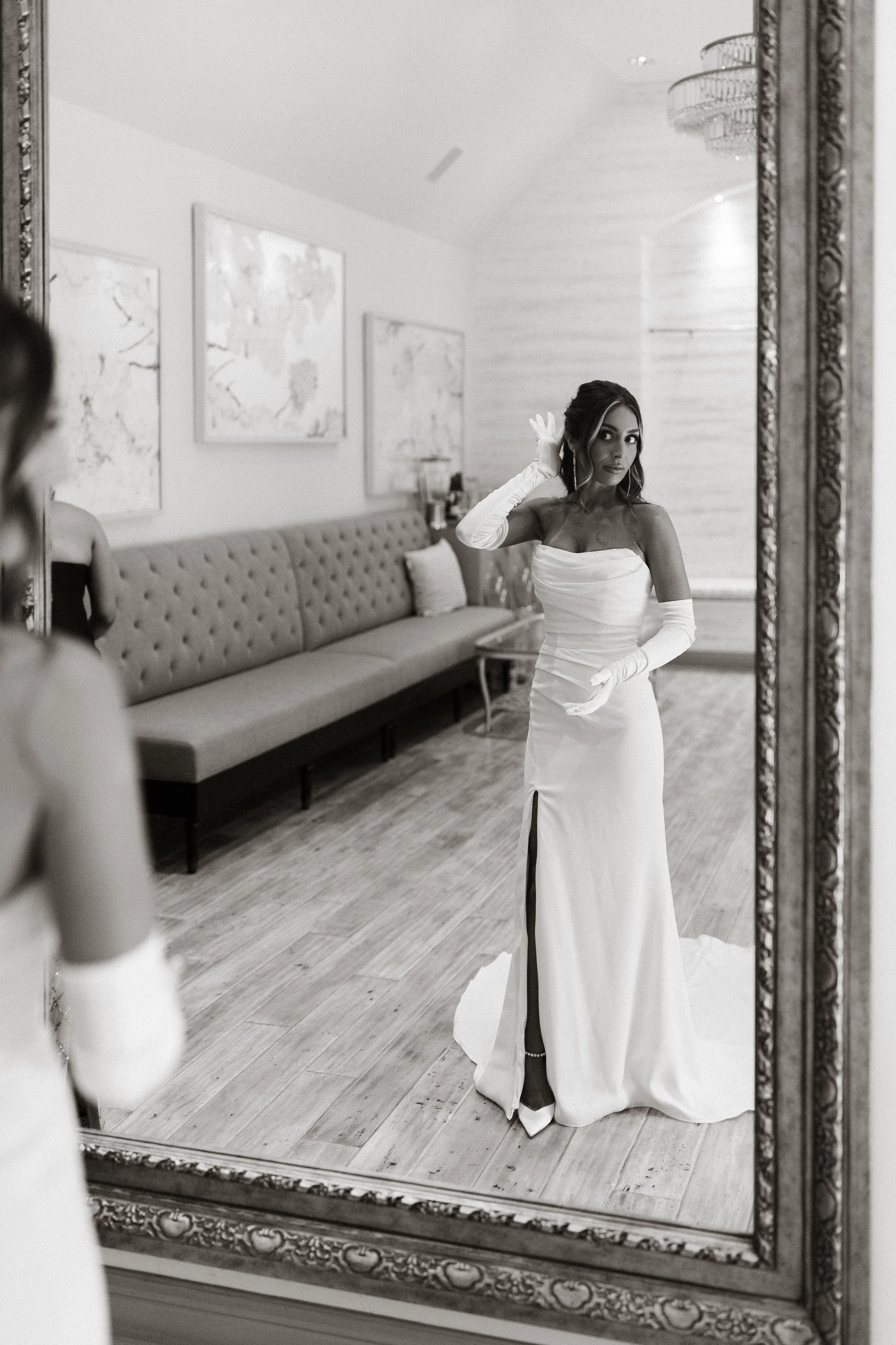 A woman in a wedding dress with long gloves is taking a mirror selfie in a room.