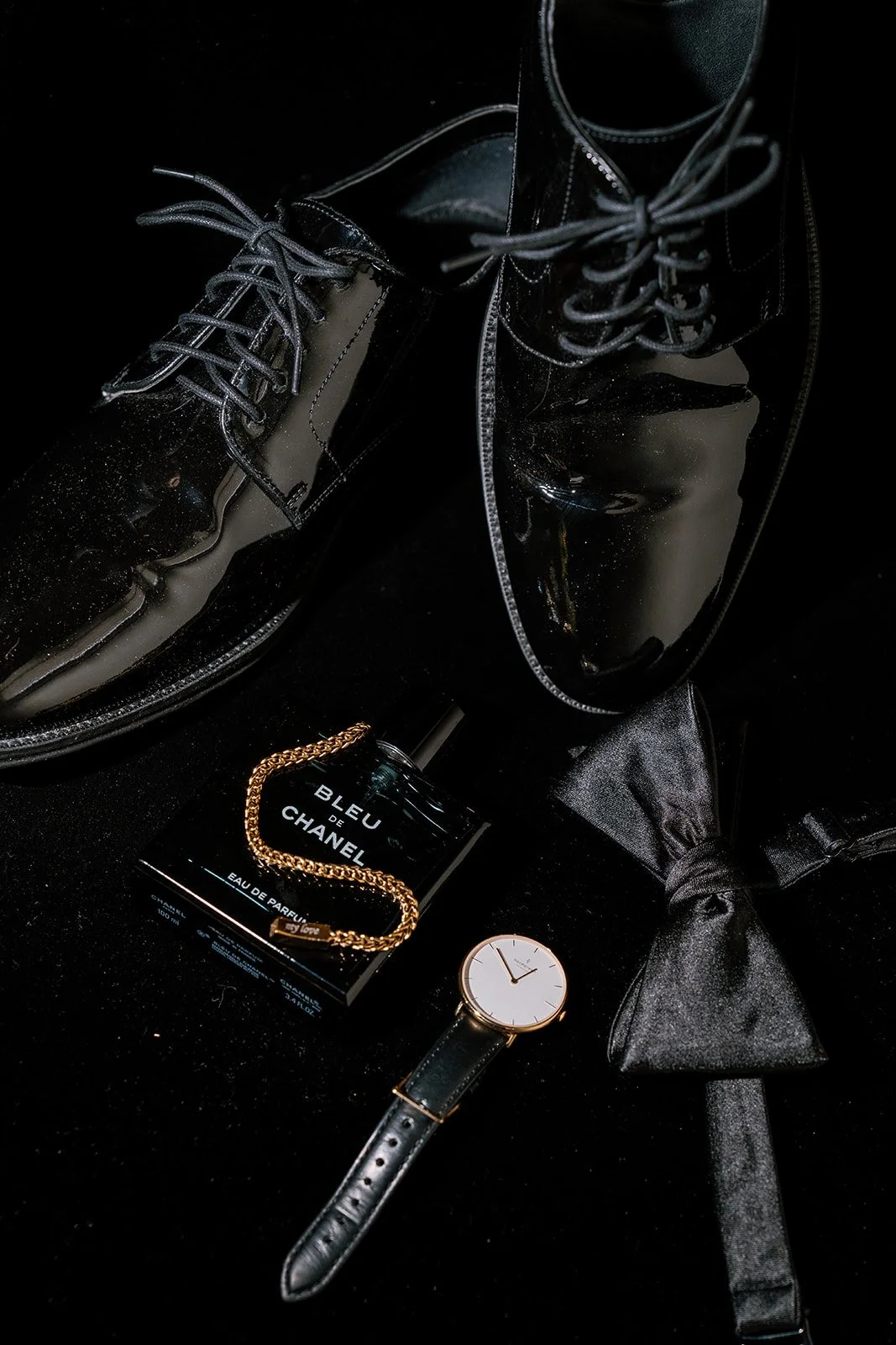 Black shiny boots, a bottle of Bleu de Chanel perfume, a black leather watch with a white face, and a black bow tie on a dark surface.