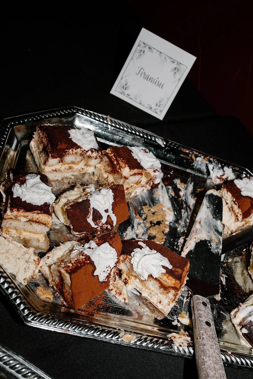 Slices of tiramisu dessert on a silver tray, with a sign labeled 'Tiramisu' in the background.