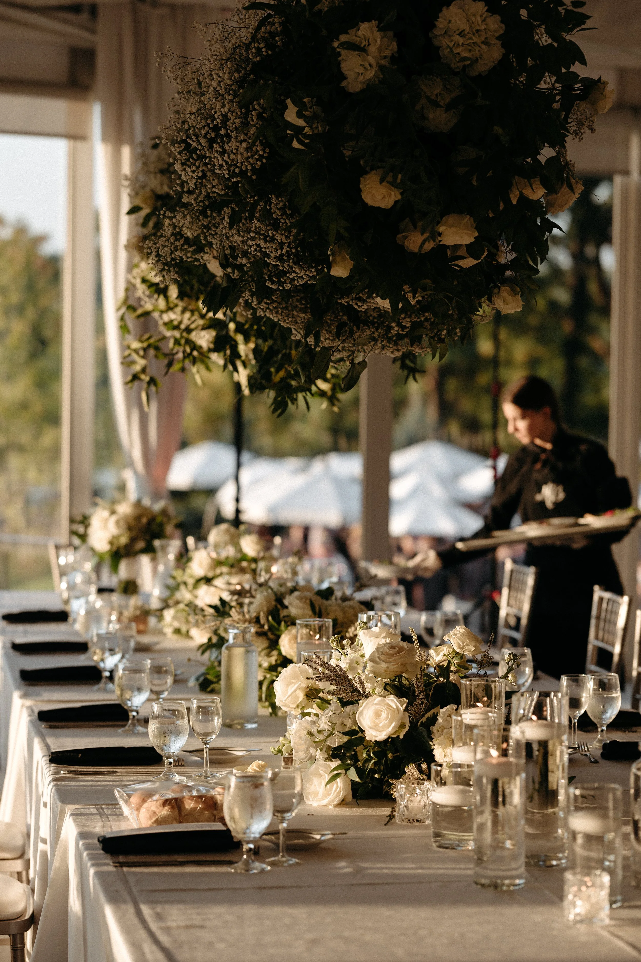 Elegant banquet table with white floral centerpieces, glassware, and black napkins under a hanging floral arrangement in a bright outdoor setting.