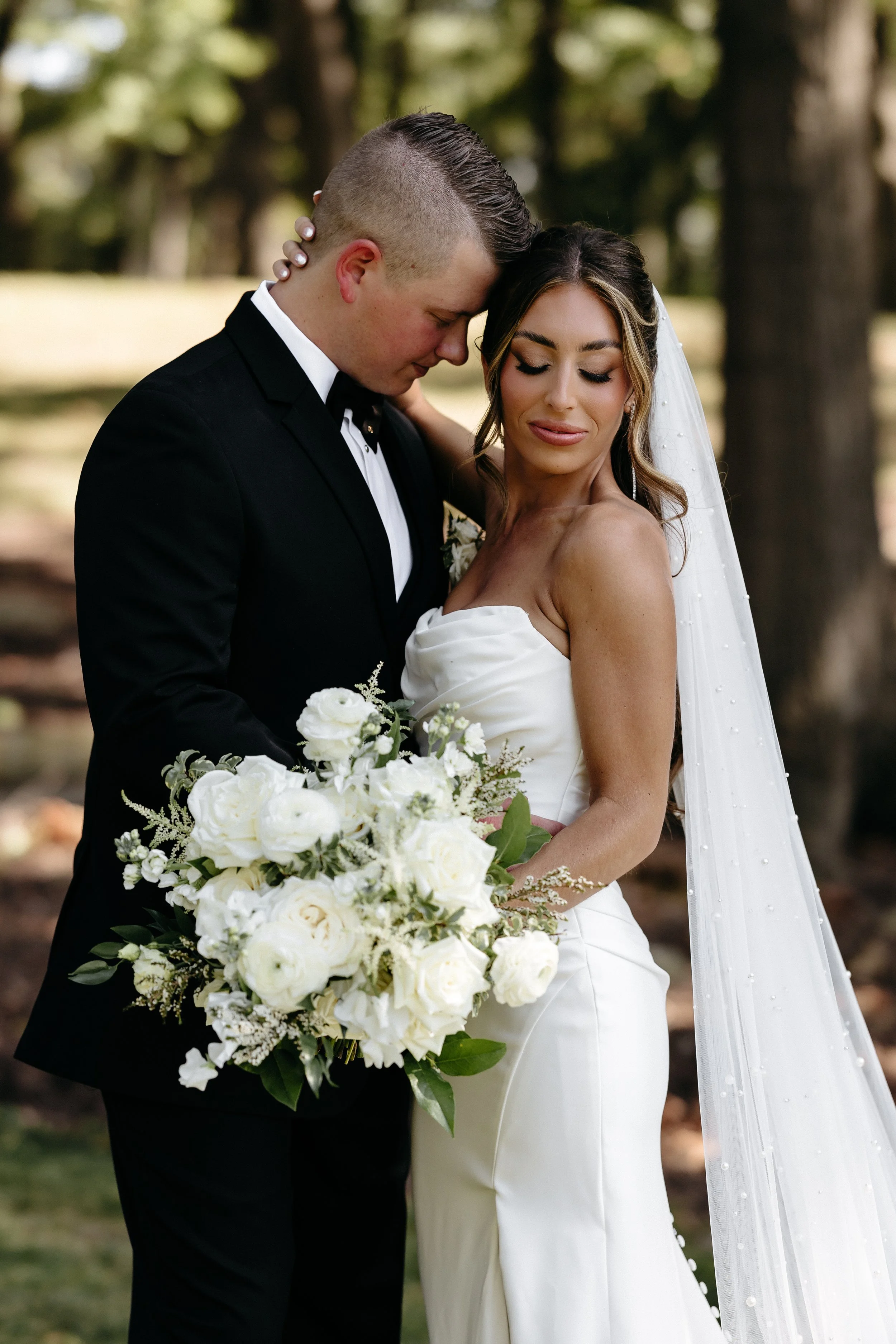 Bride and groom embracing outdoors, the bride holding a bouquet of white flowers, the groom wearing a black tuxedo, trees in the background.