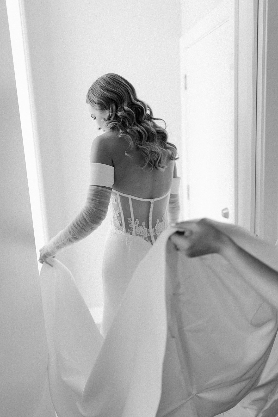 A woman in a wedding dress with lace details, wearing long gloves, is holding the fabric of her gown while looking down, in a softly lit room.