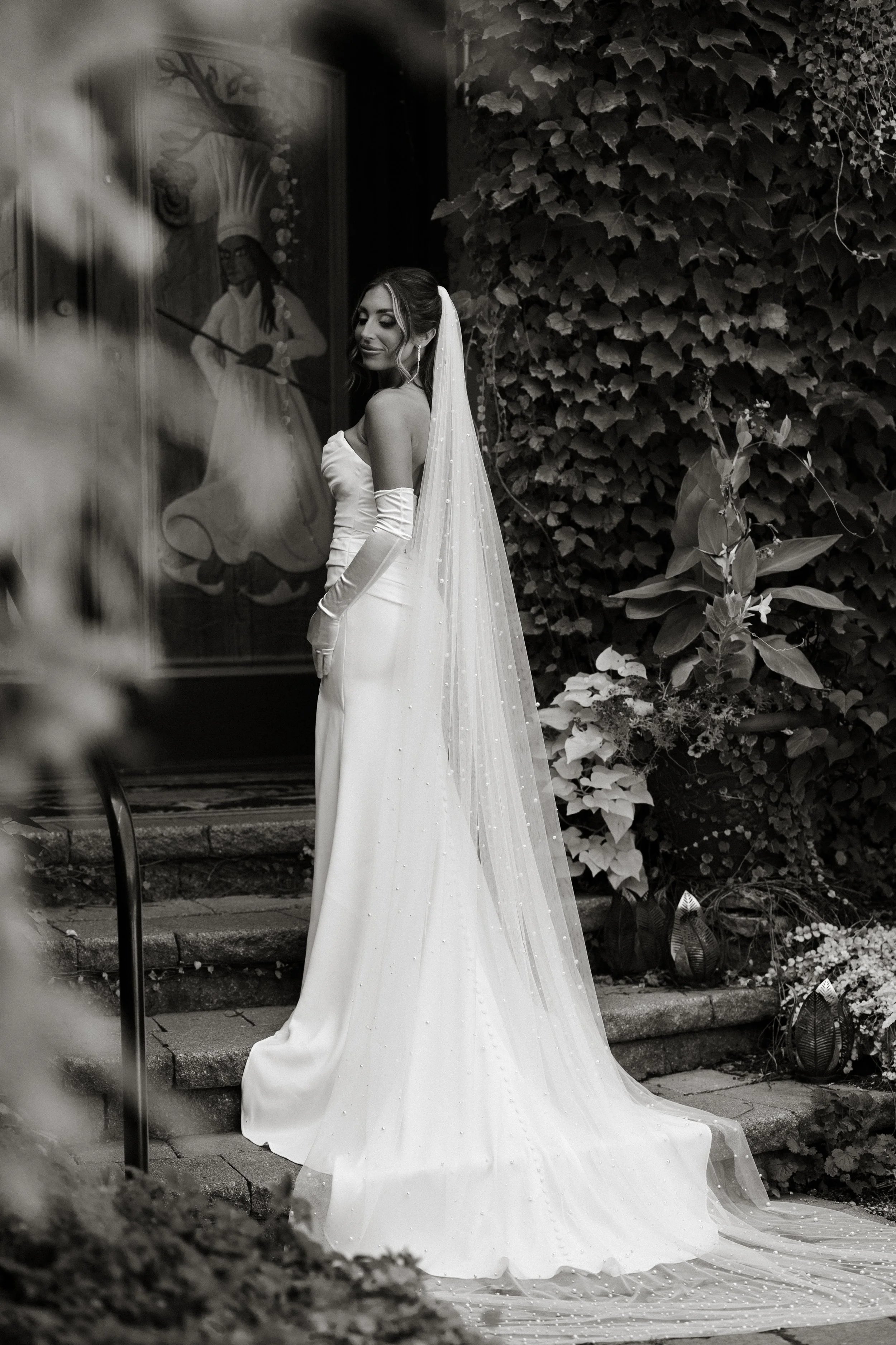 A woman in a wedding dress with long gloves and a veil, standing on steps outside near greenery and a religious painting.