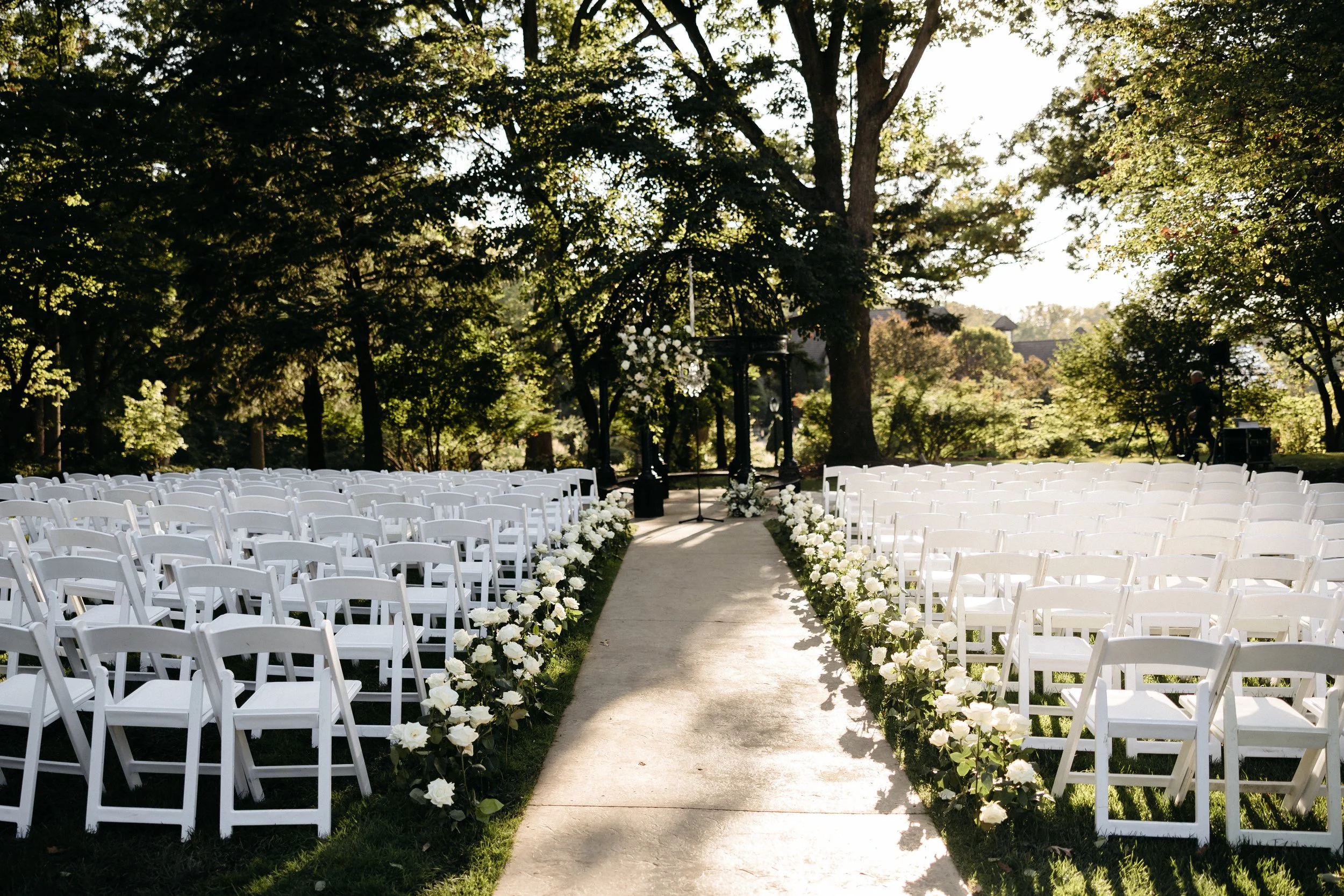 Outdoor wedding ceremony setup with white folding chairs arranged on both sides of a central aisle decorated with white roses, in a park-like setting with trees and sunlight.