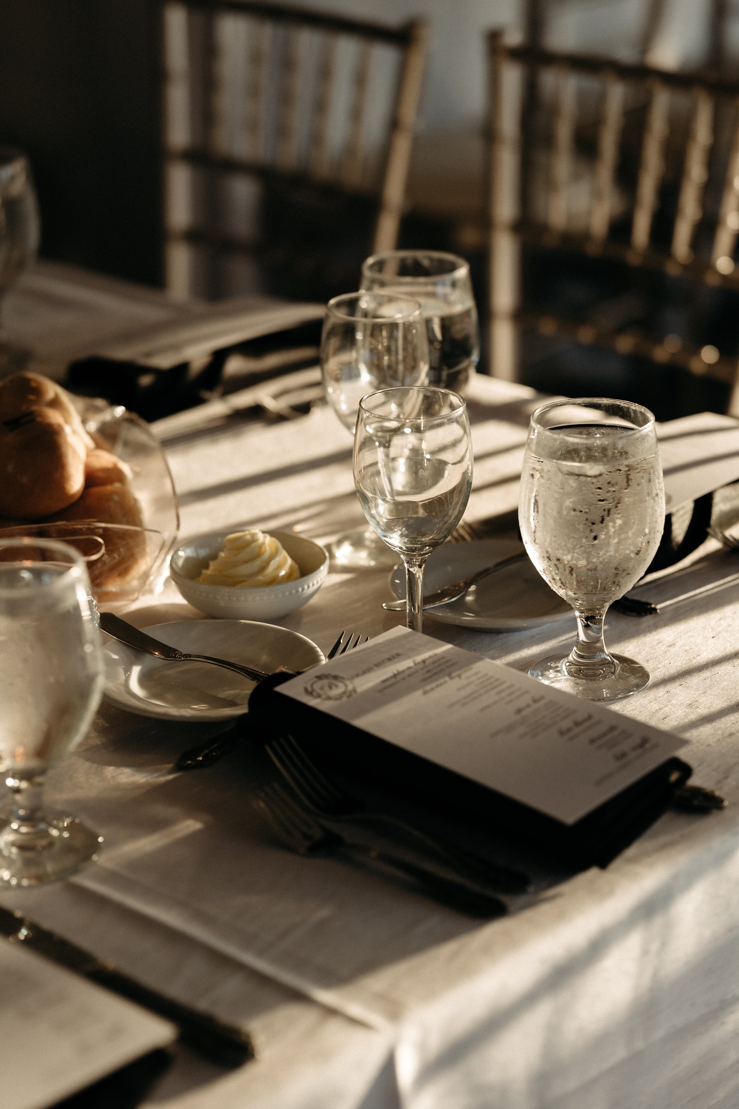 A dining table set with water glasses, a bowl of butter, bread rolls, utensils, and a menu, with sunlight casting shadows.