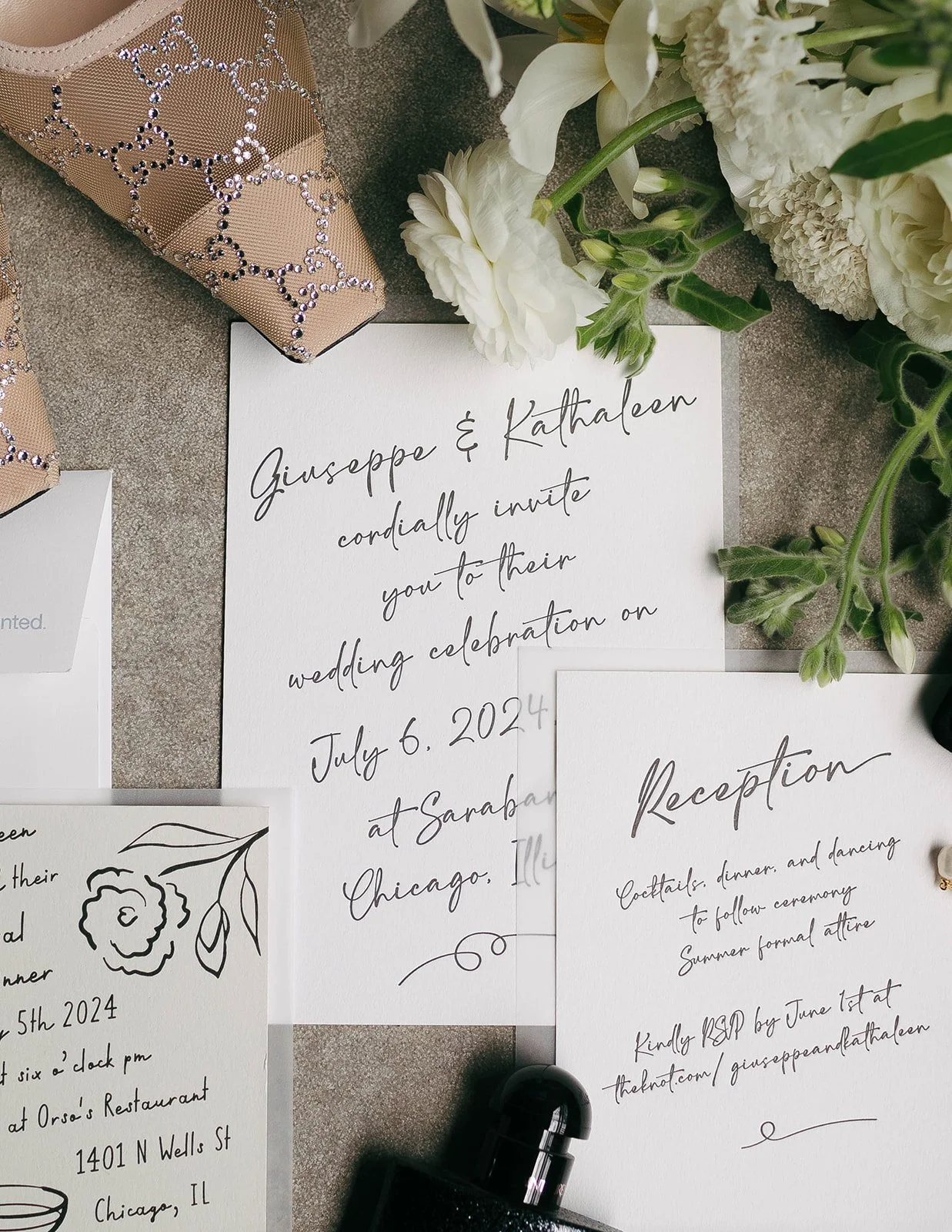 Wedding invitation cards with floral accents, a perfume bottle, and decorative shoes with rhinestones, all arranged on a textured surface.