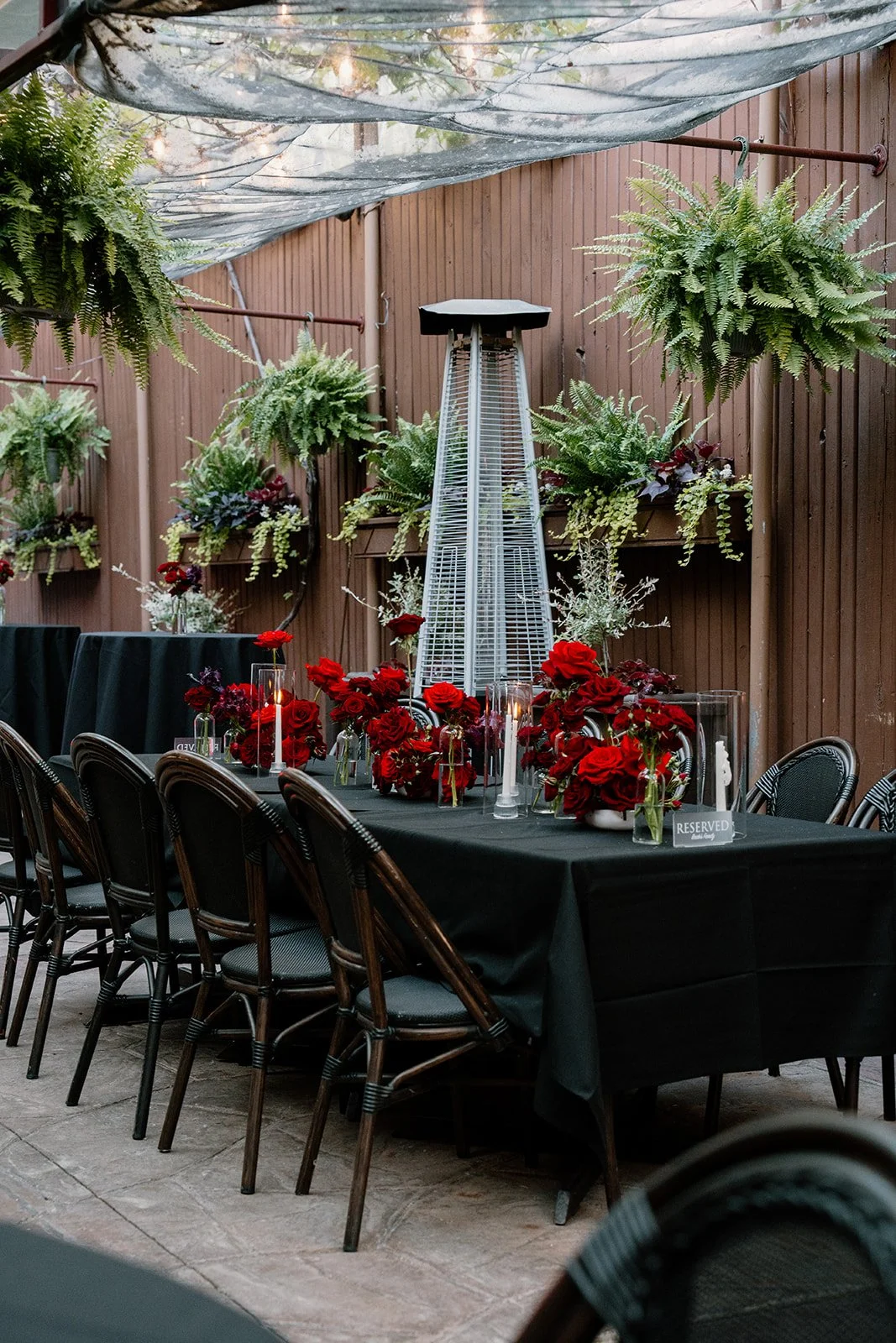 A decorated outdoor dining table with black tablecloth, red floral arrangements, and candles, set against a wooden fence with hanging fern plants and a patio heater.