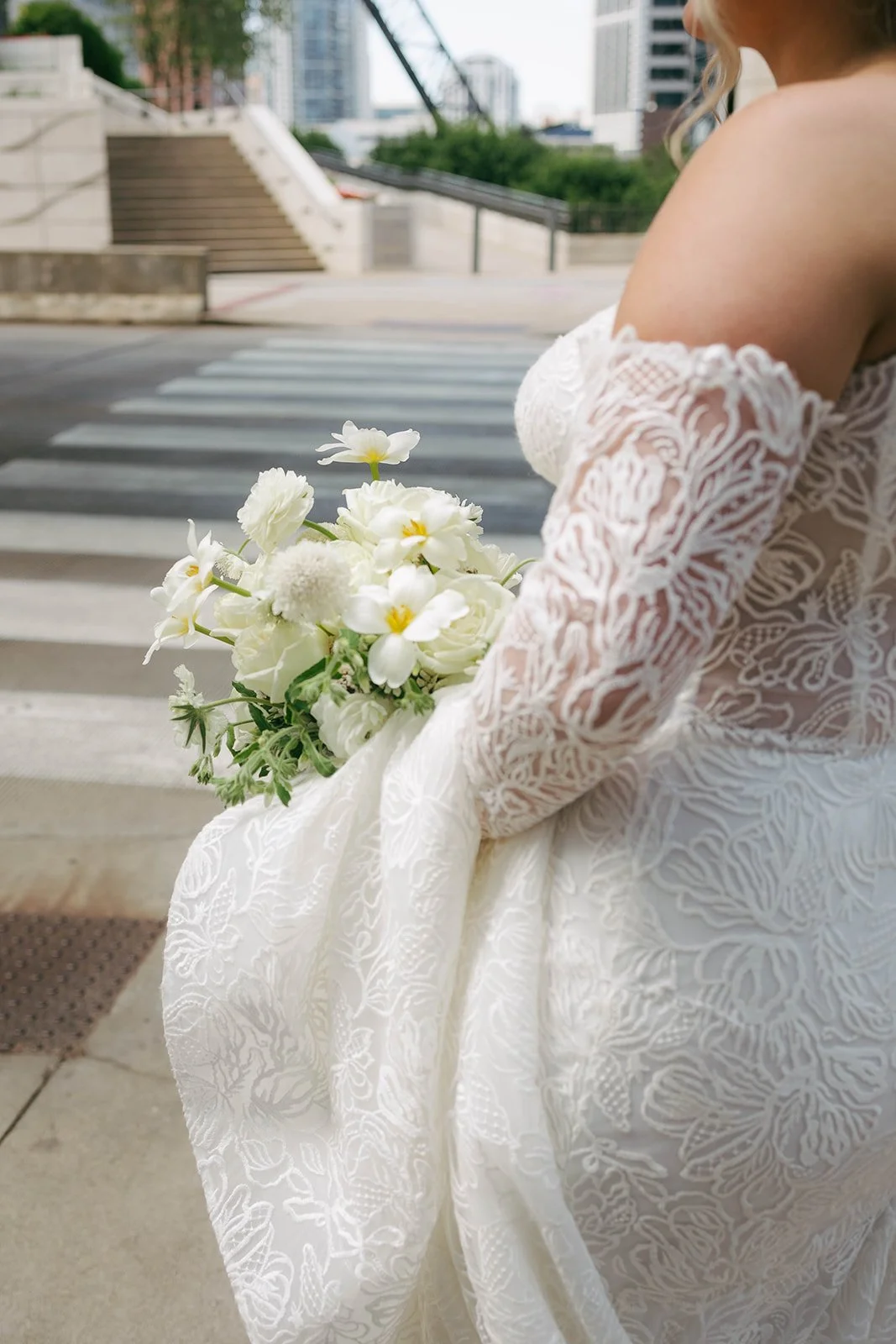 A woman in a white lace off-shoulder dress holding a bouquet of white flowers near a crosswalk in an urban setting.