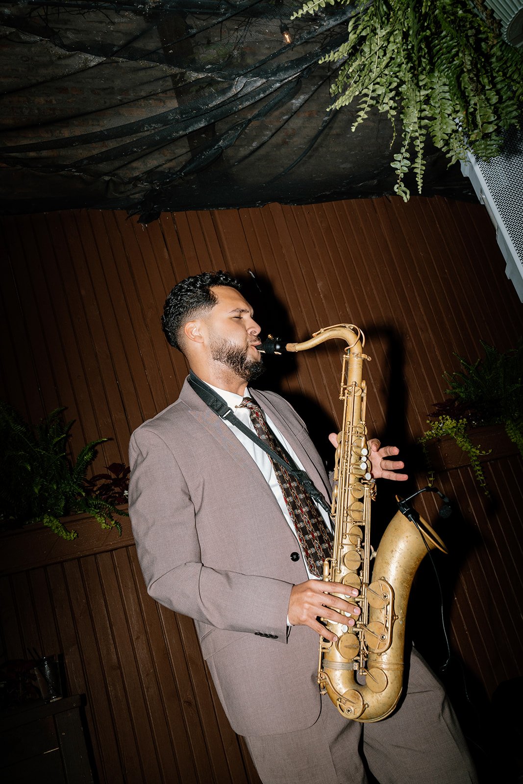 A man in a gray suit playing a saxophone with his eyes closed, standing indoors against a wood-paneled wall with some green plants.