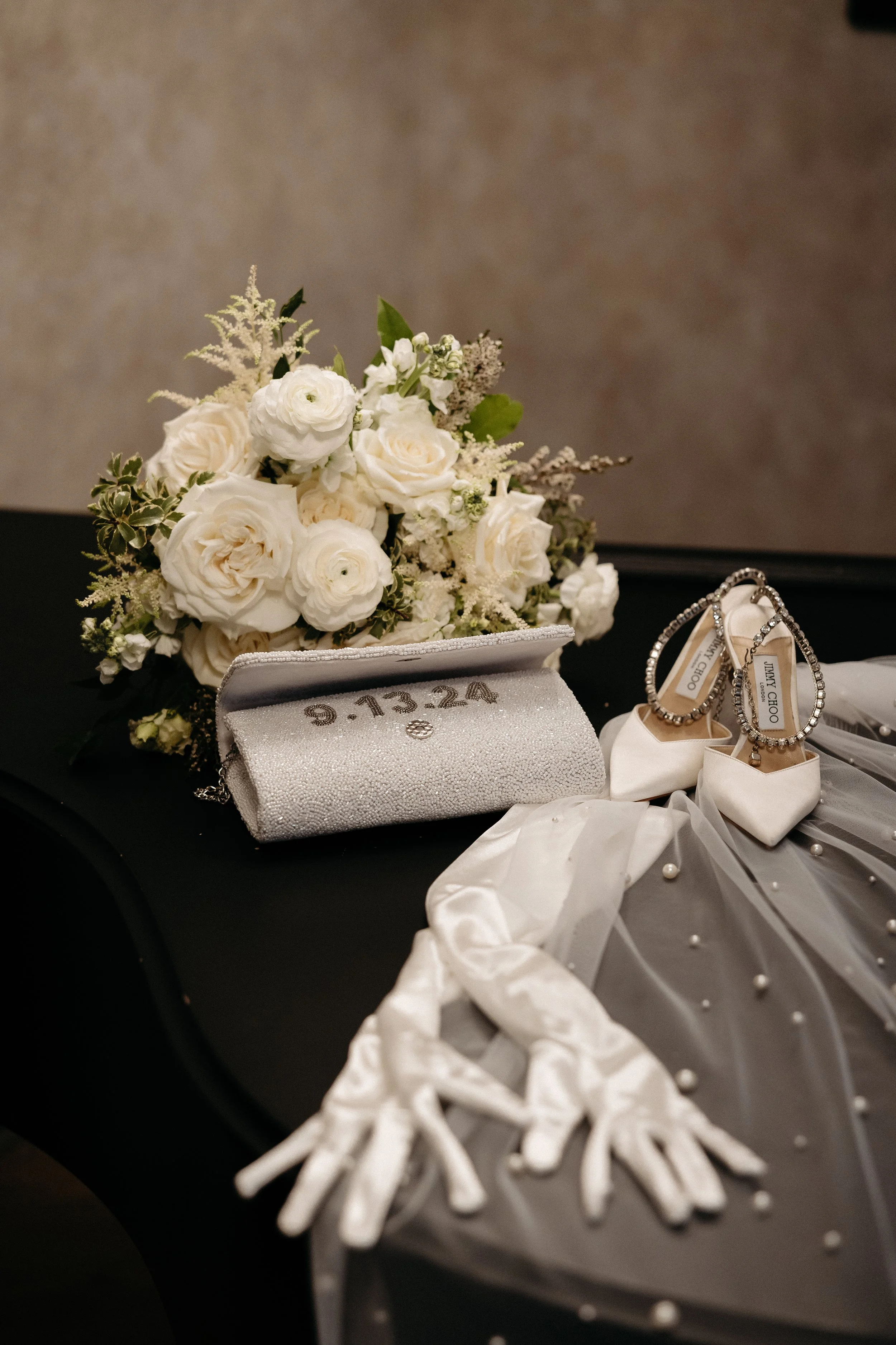 White floral bouquet with roses and greenery, silver clutch purse with date embroidery, white bridal shoes, pearl earrings, and silver bracelet on a black surface.