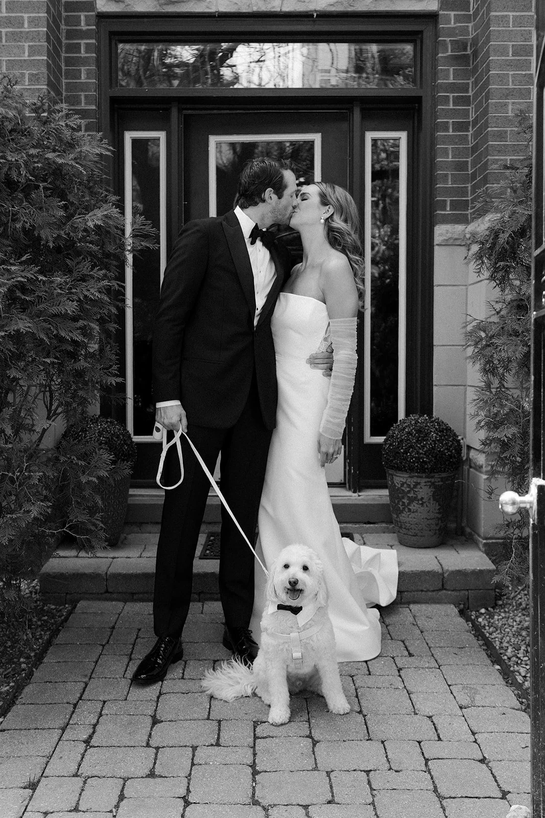 Black and white photo of a newlywed couple kissing on the front doorstep, with a dog on a leash sitting in front of them.