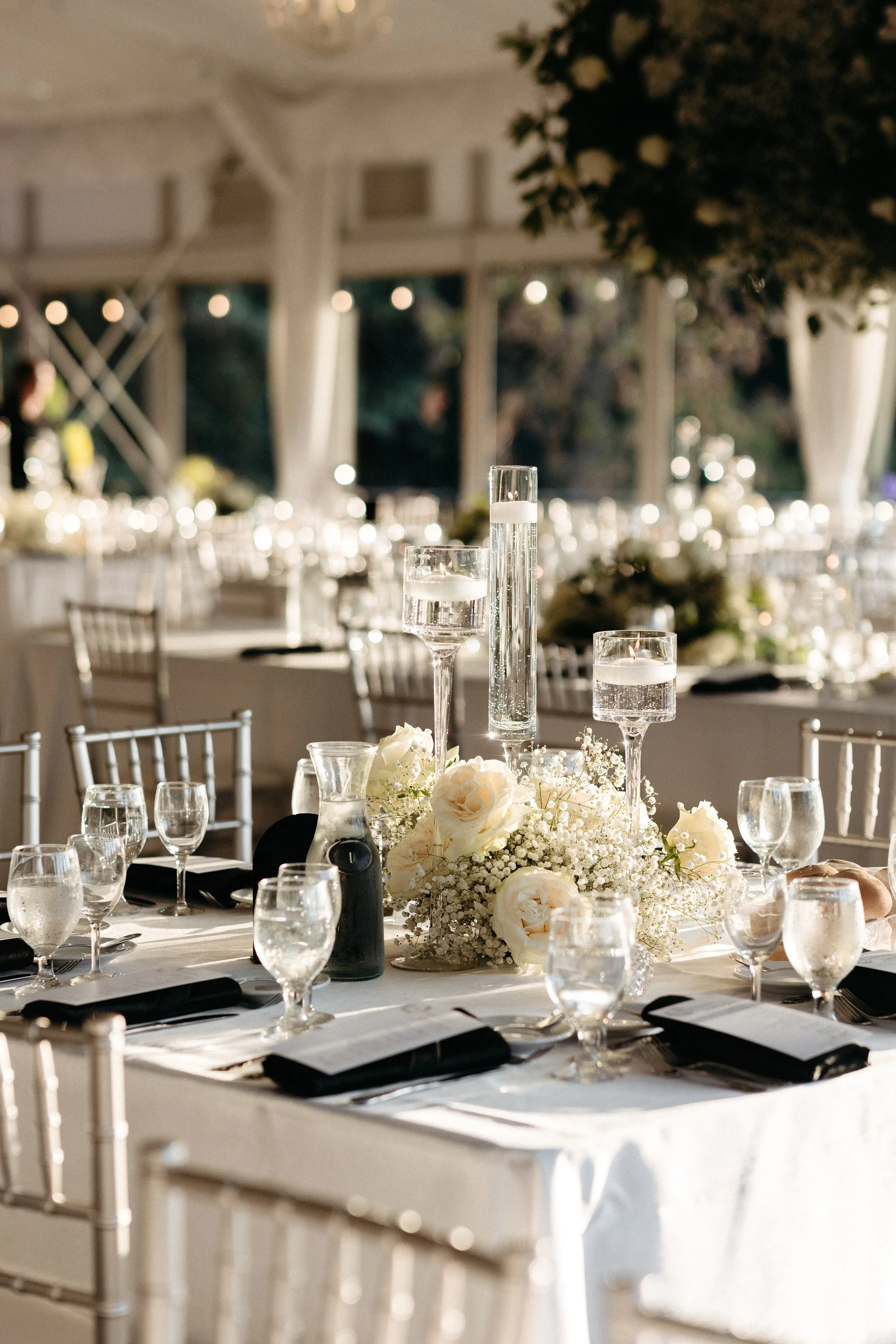 Elegant banquet table decorated with white flowers, candles in tall glass holders, and glassware, set in a decorated tent for a formal event.