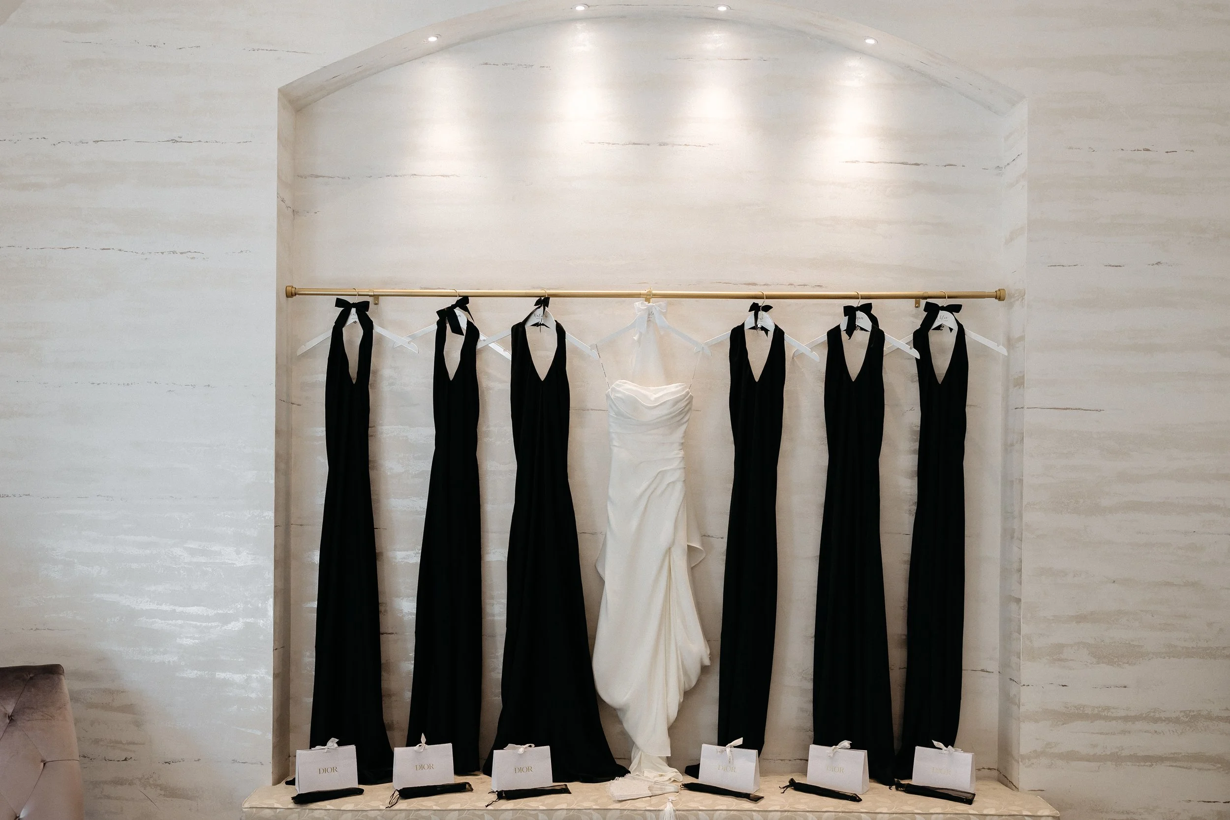 A bridal gown surrounded by six black dresses hanging on a gold rod, with gift bags labeled "Dior" at the base.