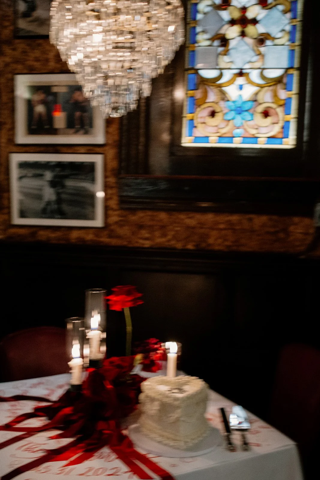 A table with a white cake, red ribbons, and red flowers, illuminated by candles in a dimly lit room with framed photos and stained glass window on the wall.