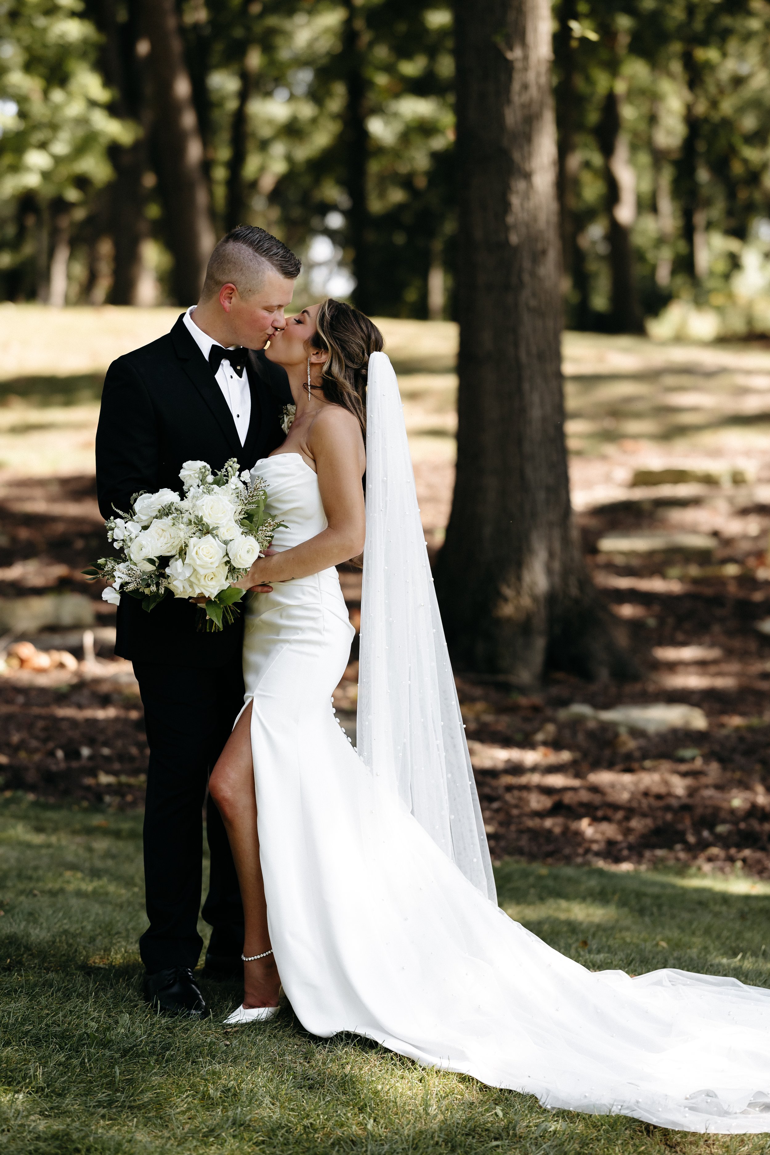 A bride and groom sharing a kiss outdoors in a wooded park, dressed in wedding attire, with the bride holding a bouquet of white roses.