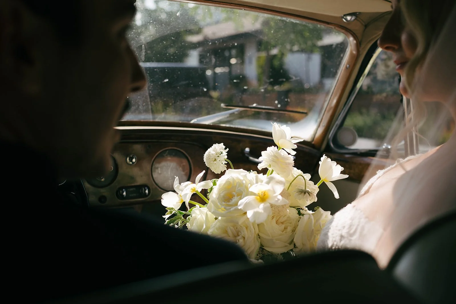A bride holding a bouquet of white flowers inside a vintage car, with a groom visible in the foreground, both facing each other.
