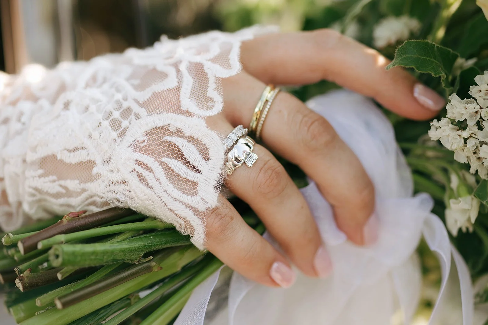 Close-up of a woman’s hand holding a bouquet of white flowers, wearing lace glove and several rings, including a skull-shaped ring with a bow and a band with diamonds.