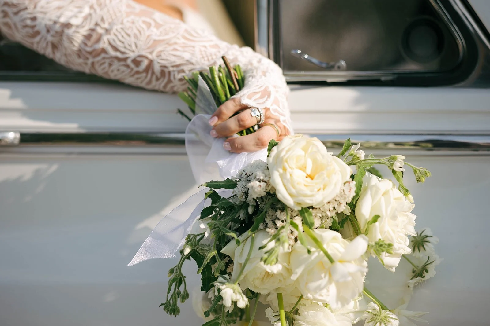 Close-up of a bride's hand holding a bouquet of white roses and greenery, with rings on her fingers, as she leans out of a vintage car window.