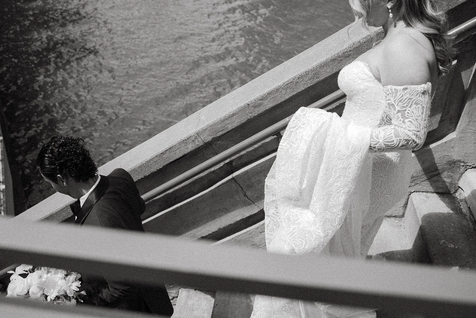A woman in a wedding dress and a man in a tuxedo sitting on stairs near a body of water, seen from above.