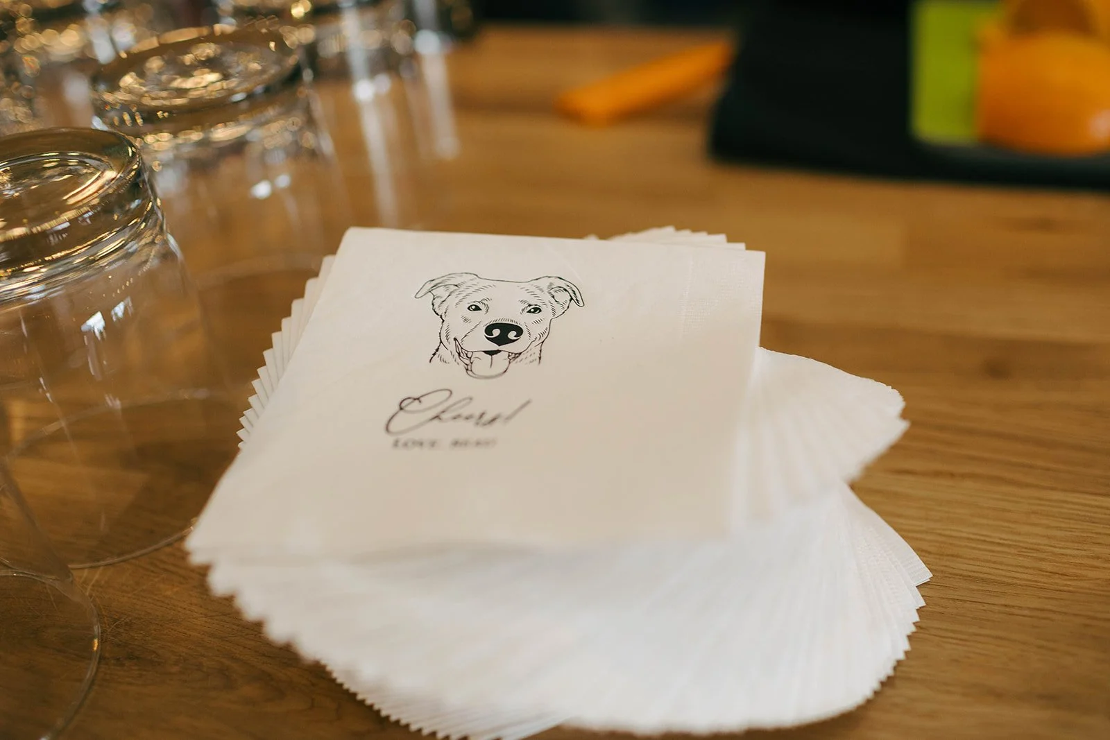 Stack of napkins with a printed dog illustration and the word "Cherish" and "Love Always" underneath, set on a wooden table with upside-down glasses and a black bag in the background.