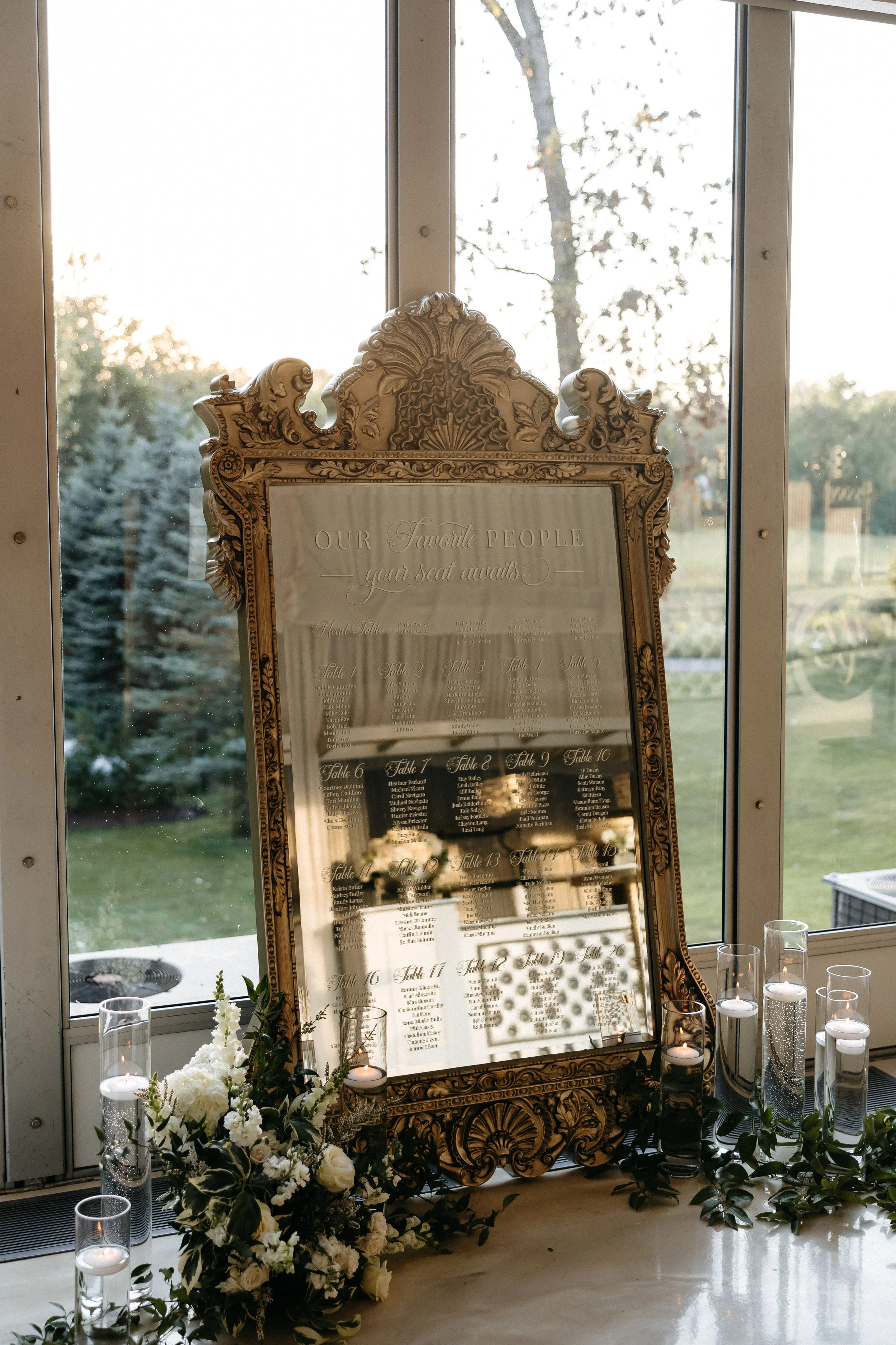 A decorative gold-framed seating chart with floral arrangements and candles, set against a large window showing trees outside.