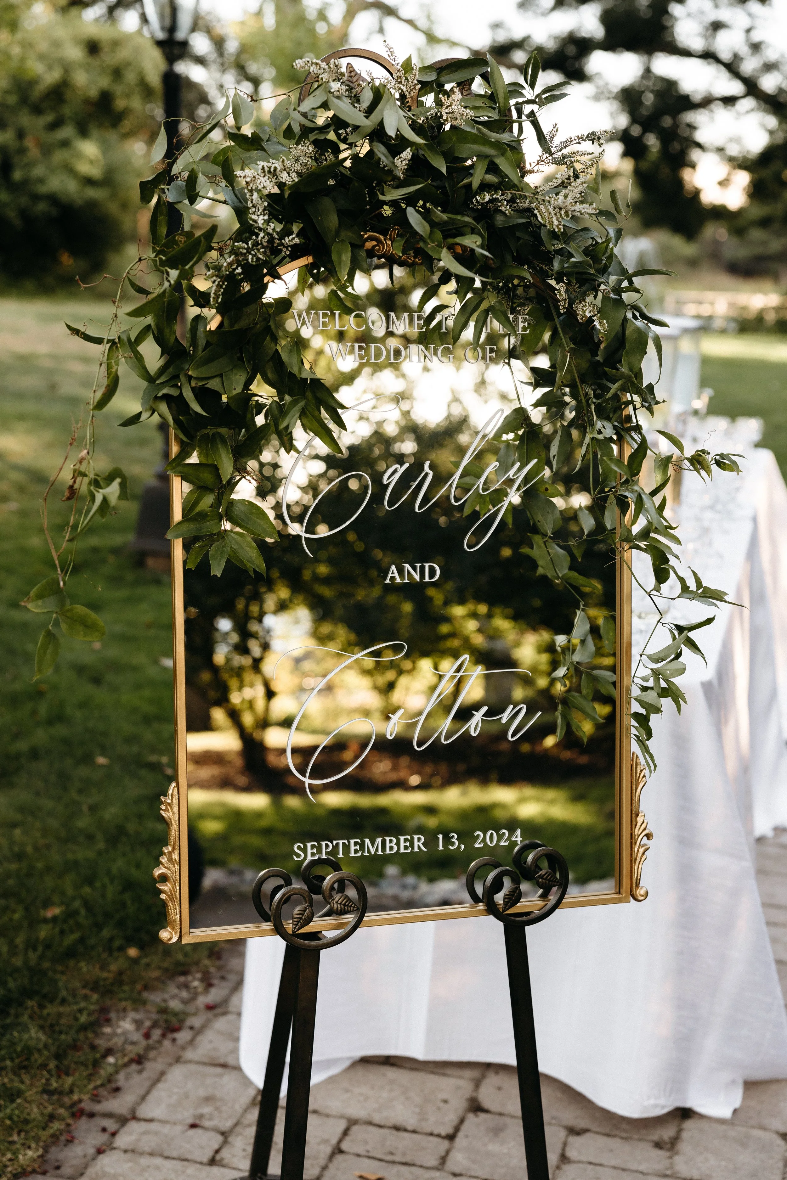 A wedding welcome sign with greenery and white flowers at the top, on a stand outdoors with trees and a white table in the background.