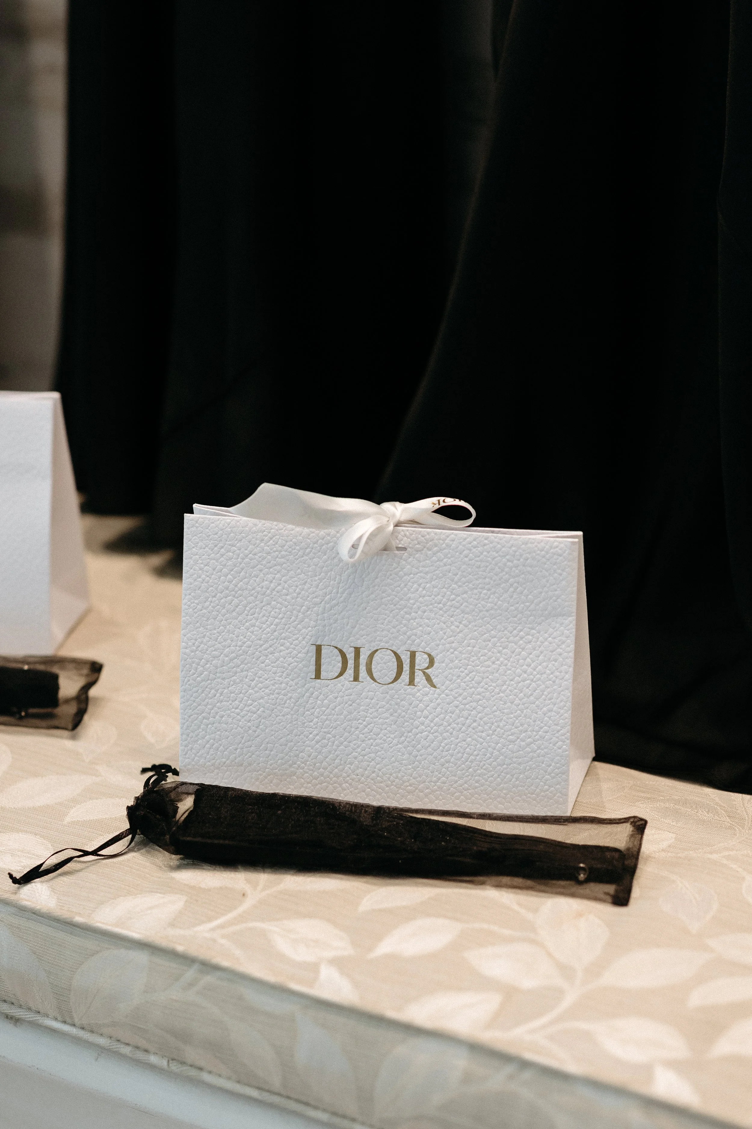 A white Dior gift bag with a textured surface and gold lettering, tied with a white ribbon, placed on a table with a beige leaf-patterned tablecloth, and a black drawstring pouch in front of it.
