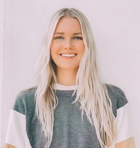 Smiling woman with shoulder-length black hair, wearing a blue top, in a green indoor setting with plants.