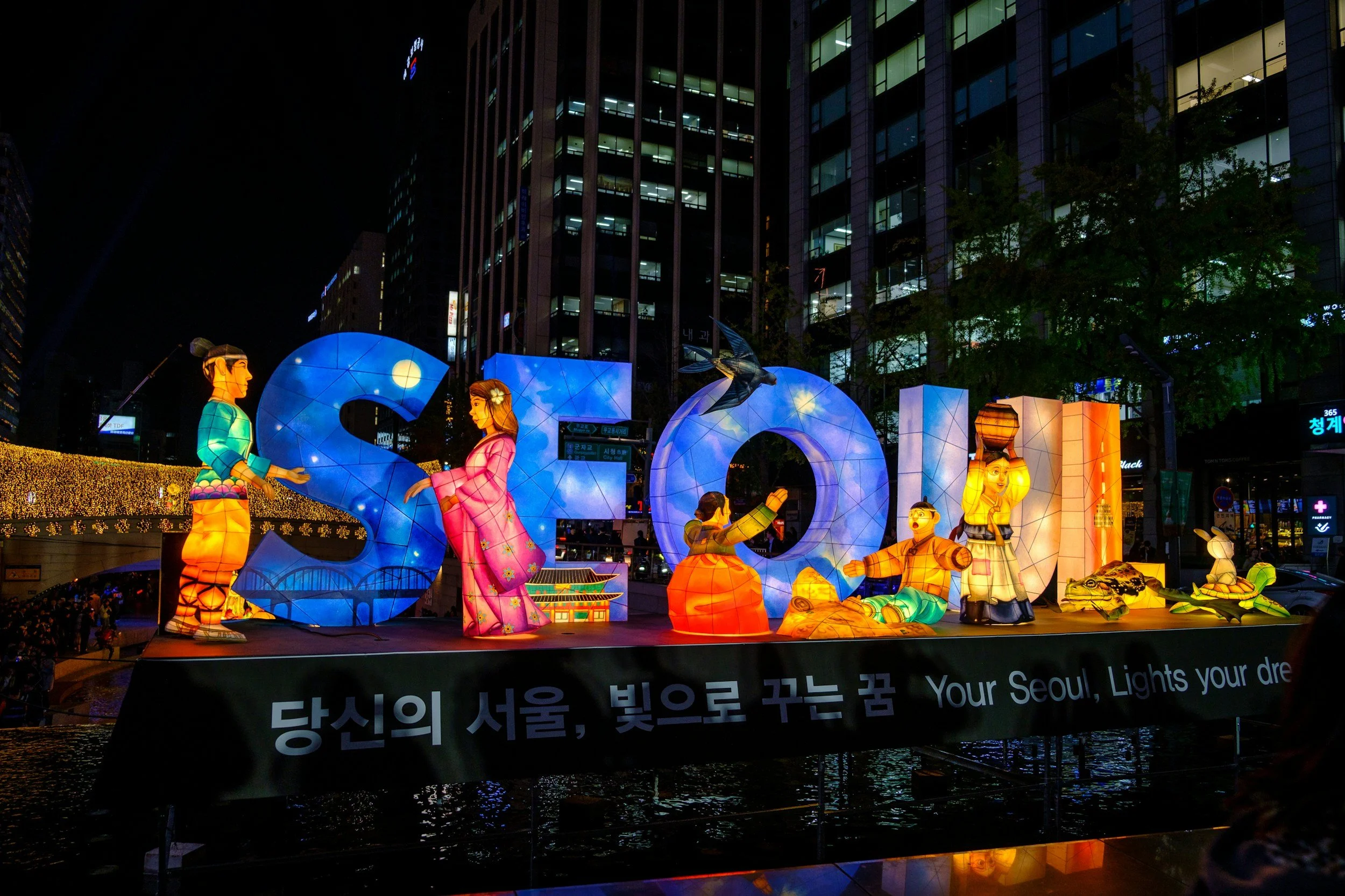 Night view of a colorful illuminated outdoor sculpture display featuring traditional Korean figures and lanterns, set against modern high-rise buildings in Seoul.