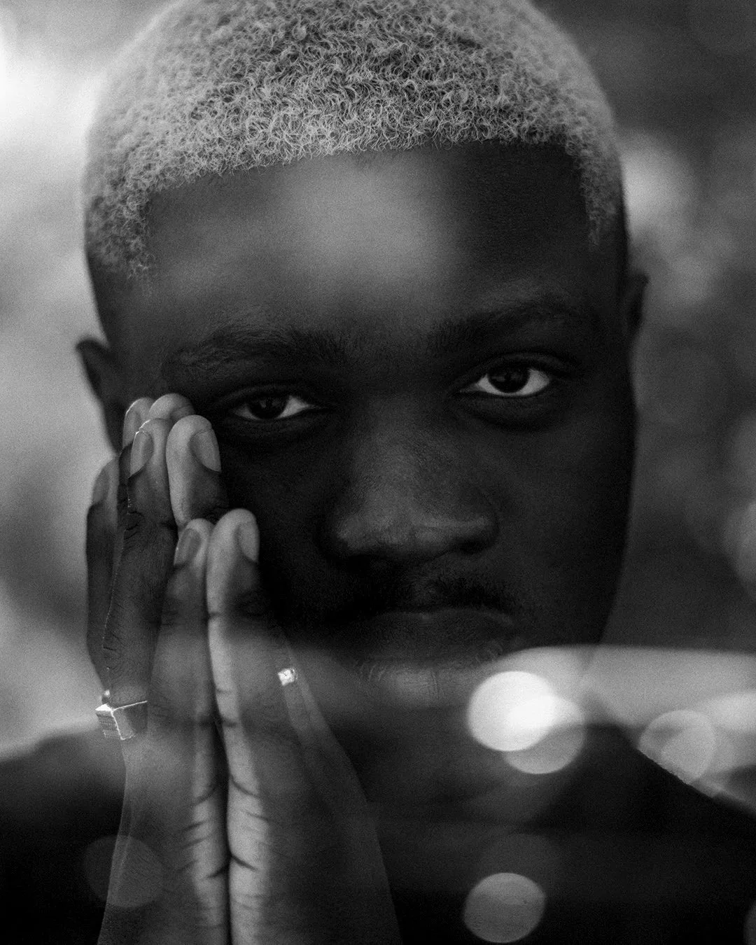 Portrait en noir et blanc d'une personne afro-américaine avec une tête rasée blonde et un regard intense, tenant sa joue avec la main gauche, portant une bague.