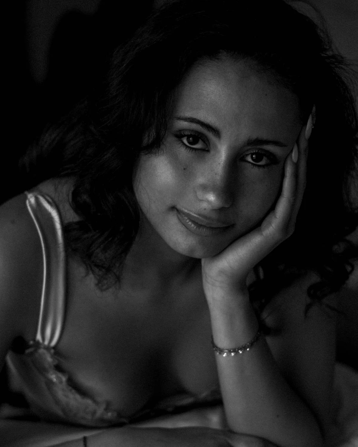 Portrait en noir et blanc d'une femme avec des cheveux bouclés, regardant vers l'objectif, posant avec la main sur la joue, portant un bracelet et un vêtement à fines bandelettes.