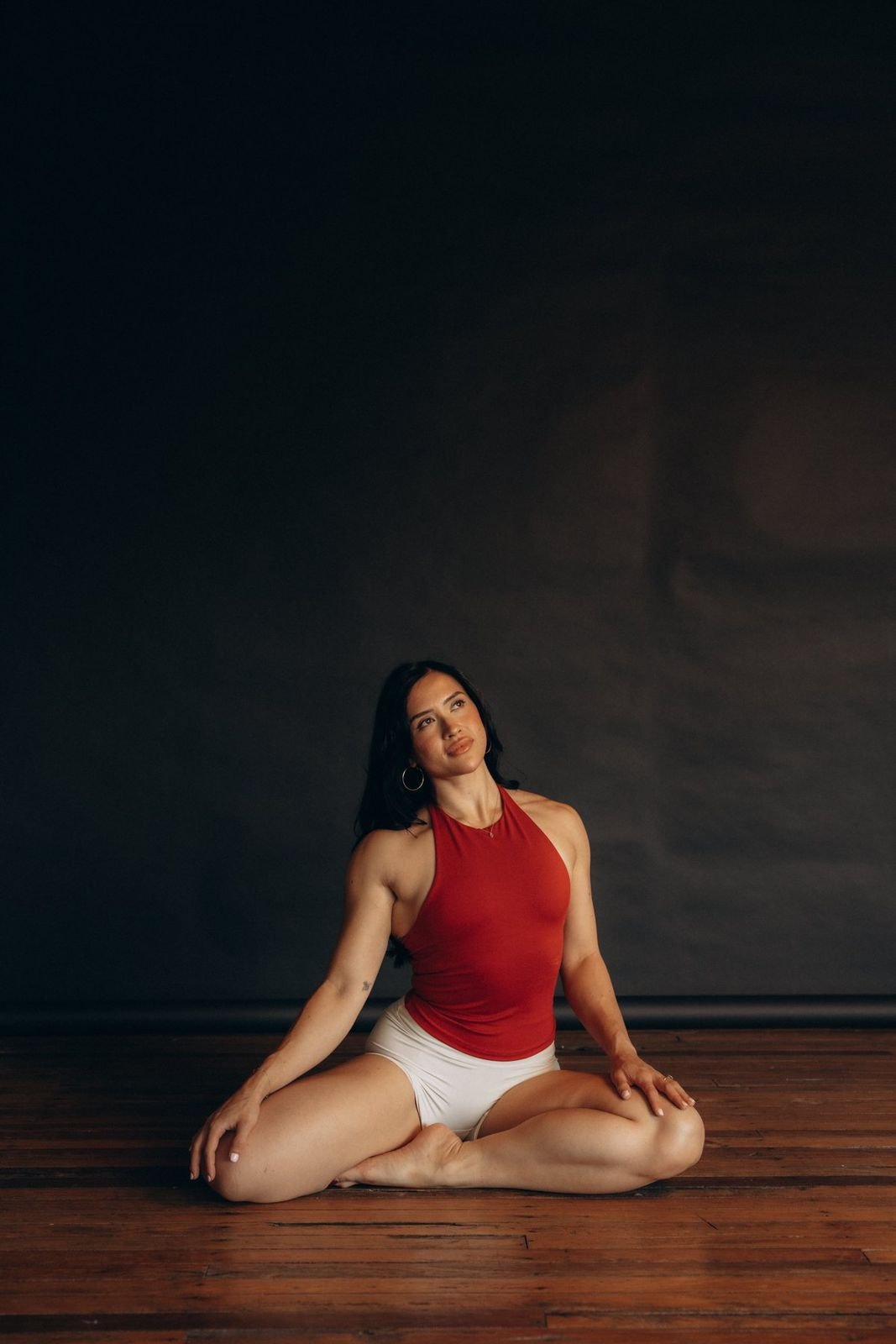 Woman in a red tank top and white shorts practicing yoga on a wooden floor in front of a dark wall.