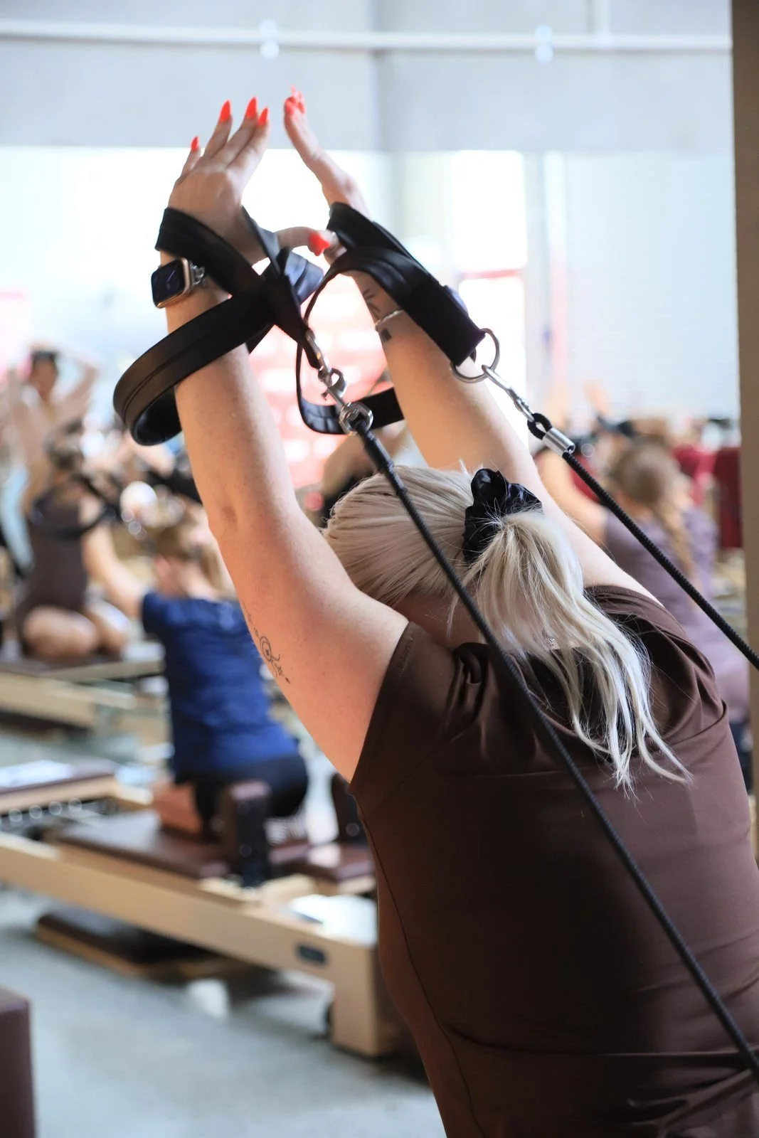 A woman with blonde hair tied back with a black hair scrunchy exercises with resistance bands, stretching her arms overhead in a fitness class.