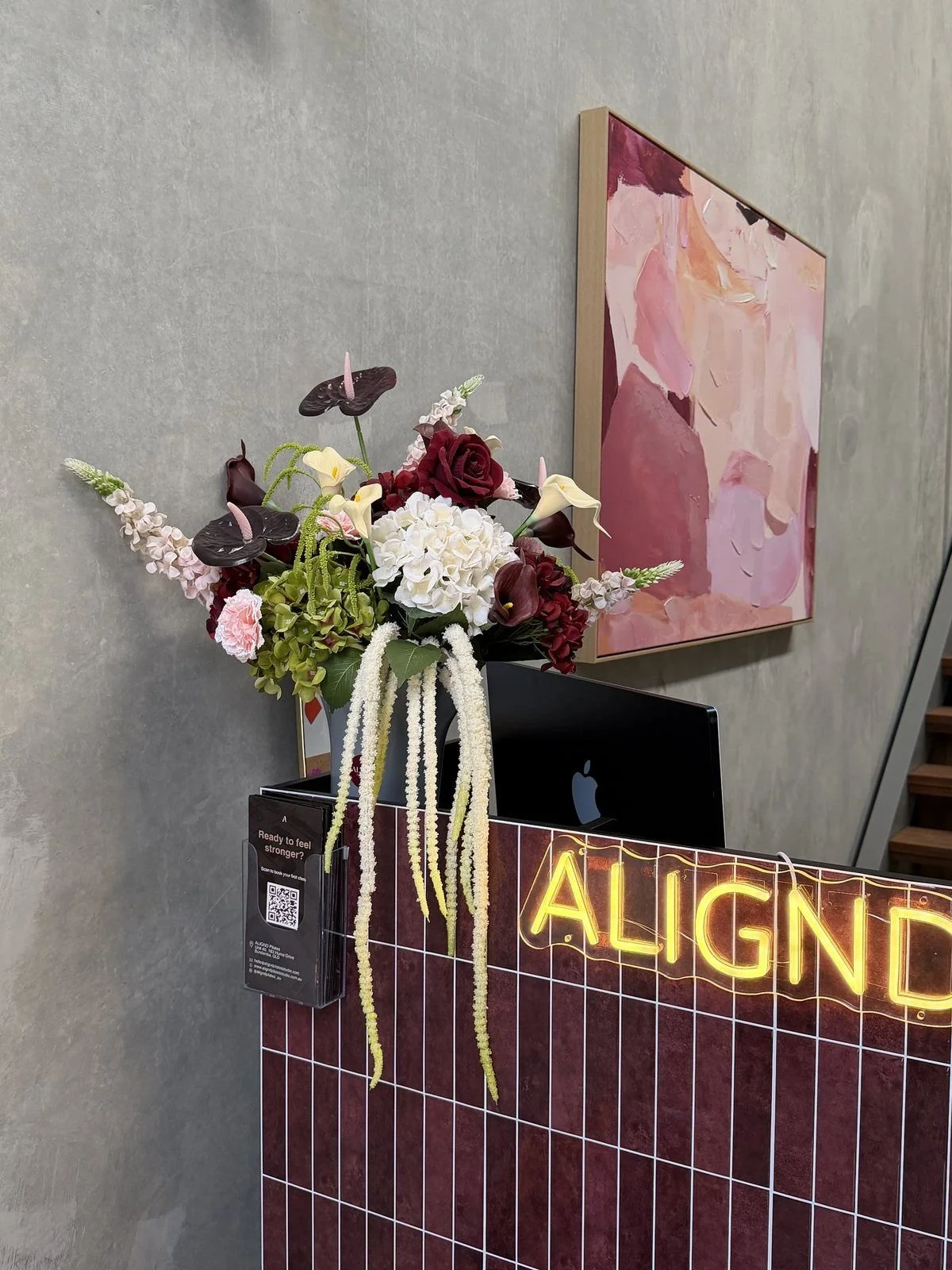 A floral arrangement featuring red roses, white hydrangeas, calla lilies, anthuriums, and pink carnations, placed on a black counter with a neon sign reading 'ALIGNED'. In the background, a colorful abstract painting hangs on the wall, and a gray wall is visible.