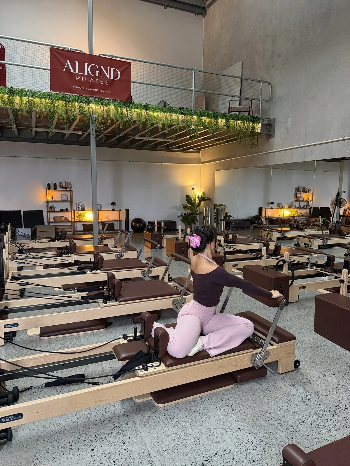 A woman practicing Pilates on a reformer machine in a fitness studio with multiple reformers, warm lighting, green plants, shelves with decor, and a mezzanine with a banner that reads 'ALIGND PILATES'.
