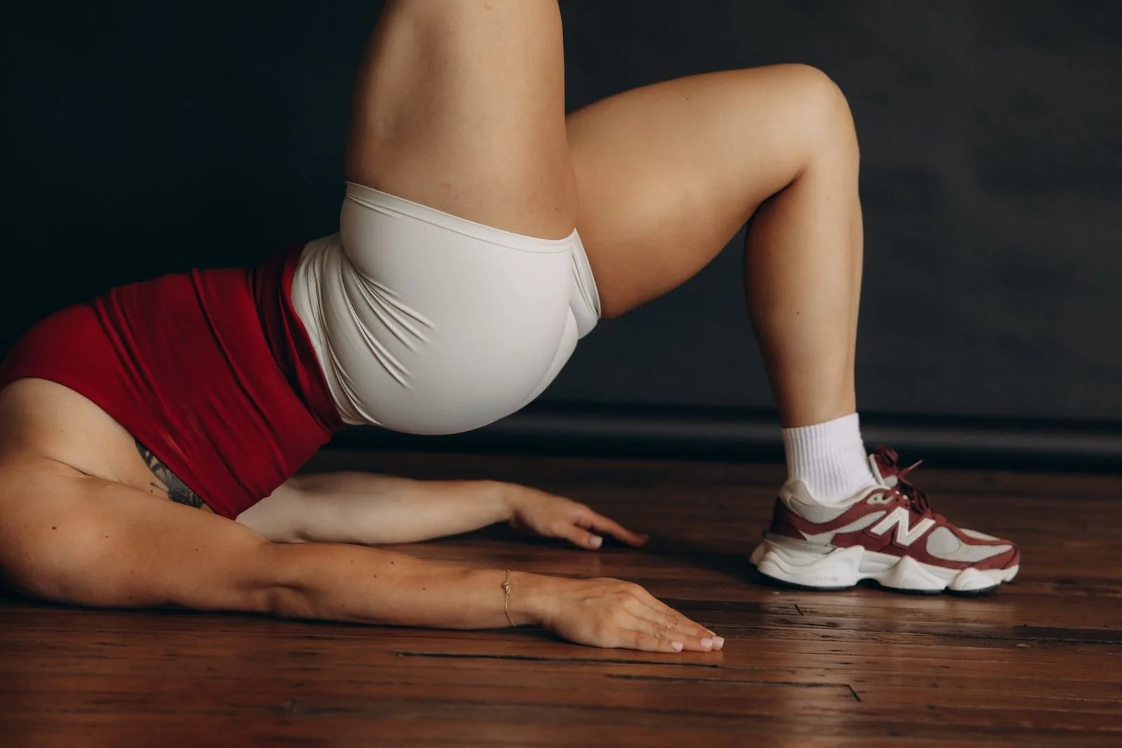A person performing a yoga pose on a wooden floor. They are wearing red and white athletic clothing and sneakers, with one leg bent and supported by their hand.