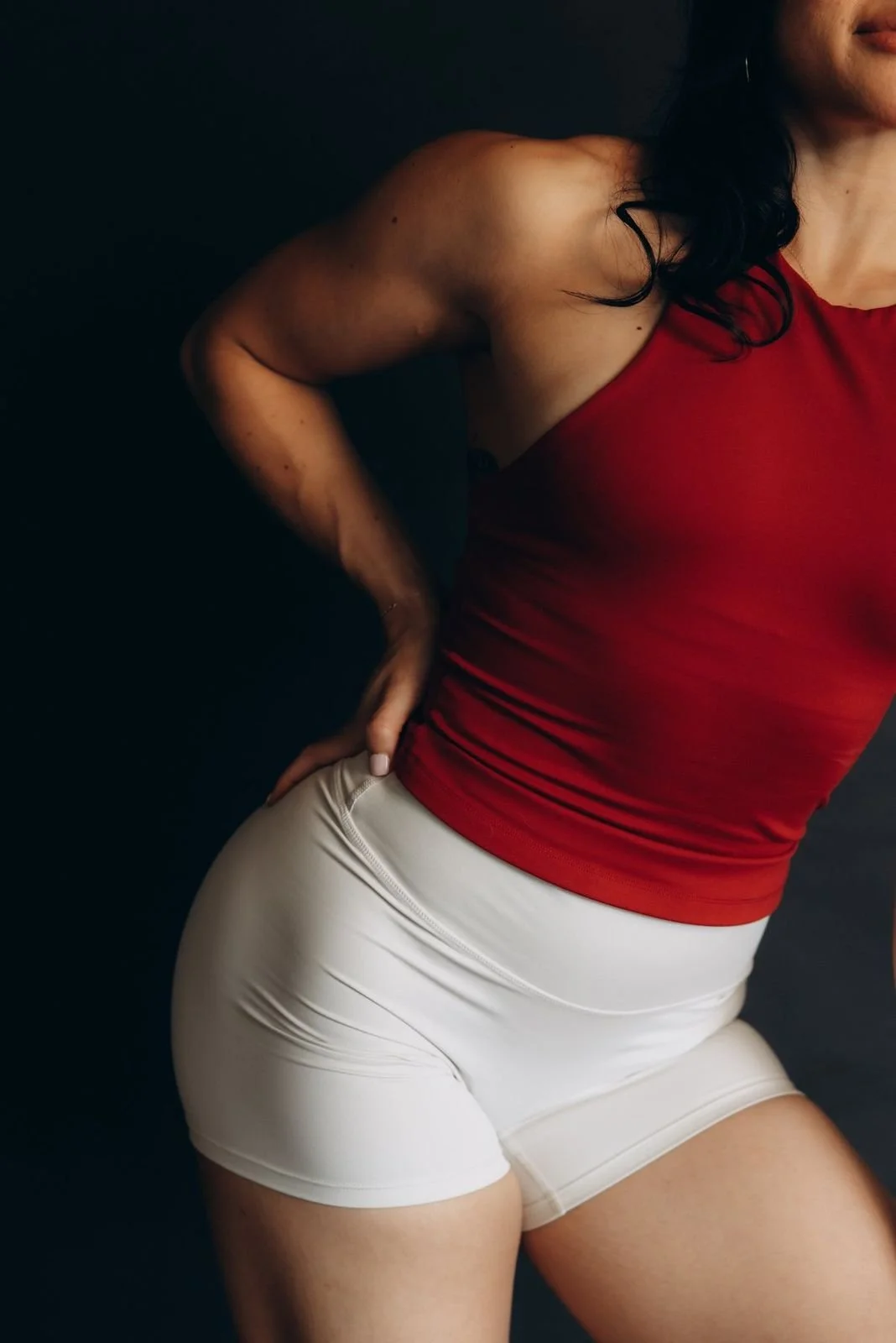 A woman wearing a red sleeveless top and beige shorts posing against a black background.