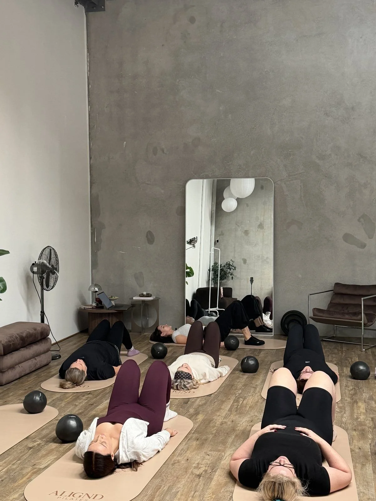 Six people lying on Pilates mats in a studio, participating in a class, with exercise balls nearby and a large mirror reflecting the room. The studio has a minimalist design with a concrete wall and wooden floor.