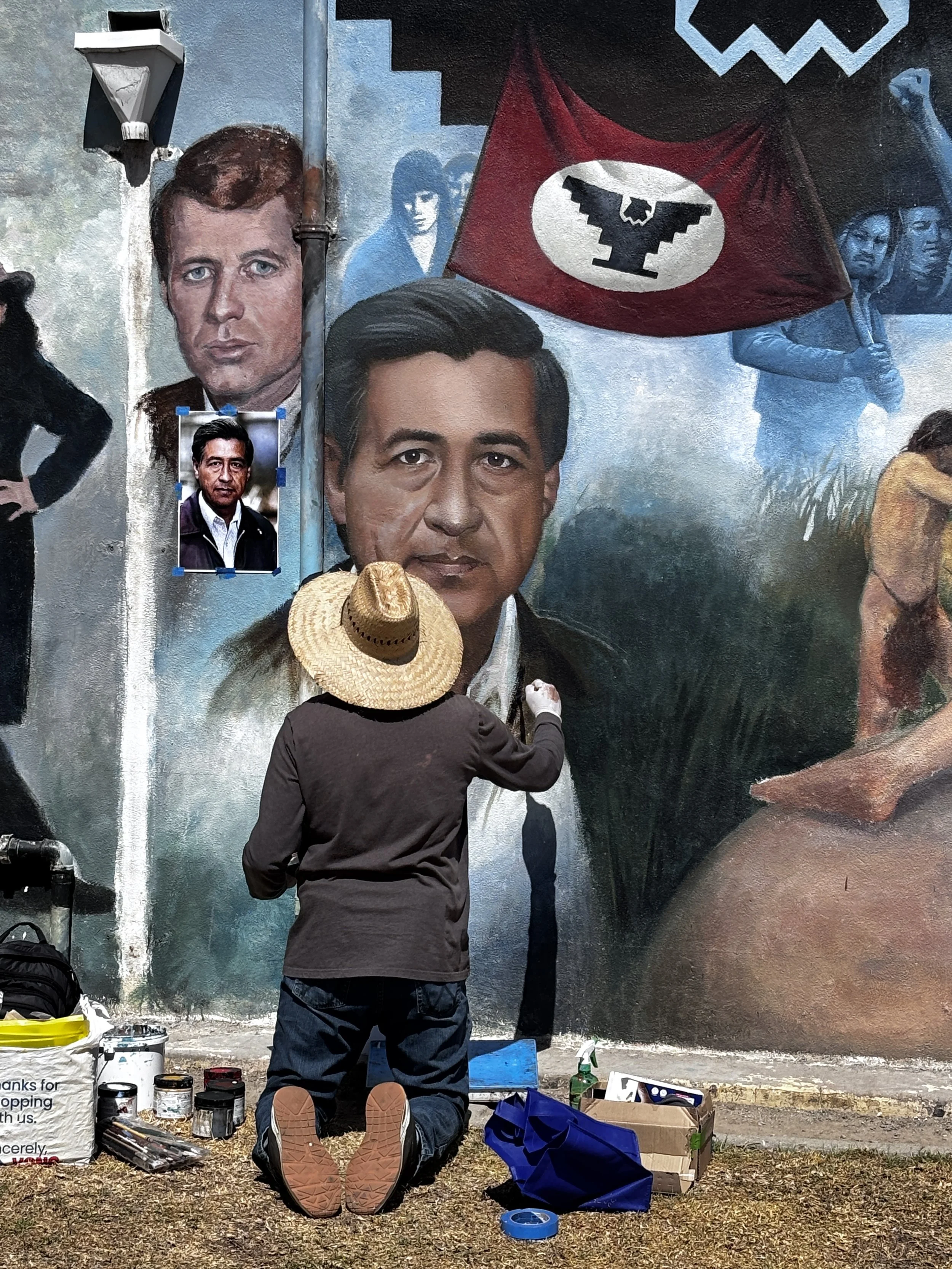 An artist painting a mural on a wall featuring portraits of individuals, including a man with dark hair and another with lighter hair, a flag with an eagle emblem, and various smaller images, with painting supplies and a person wearing a straw hat kneeling in front.
