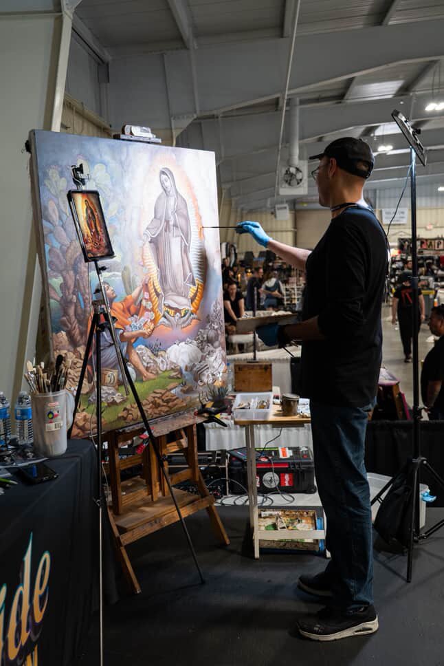 Horacio Martinez painting a religious scene on a large canvas, with religious figures and a central figure resembling the Virgin Mary, in a busy indoor market or event space.