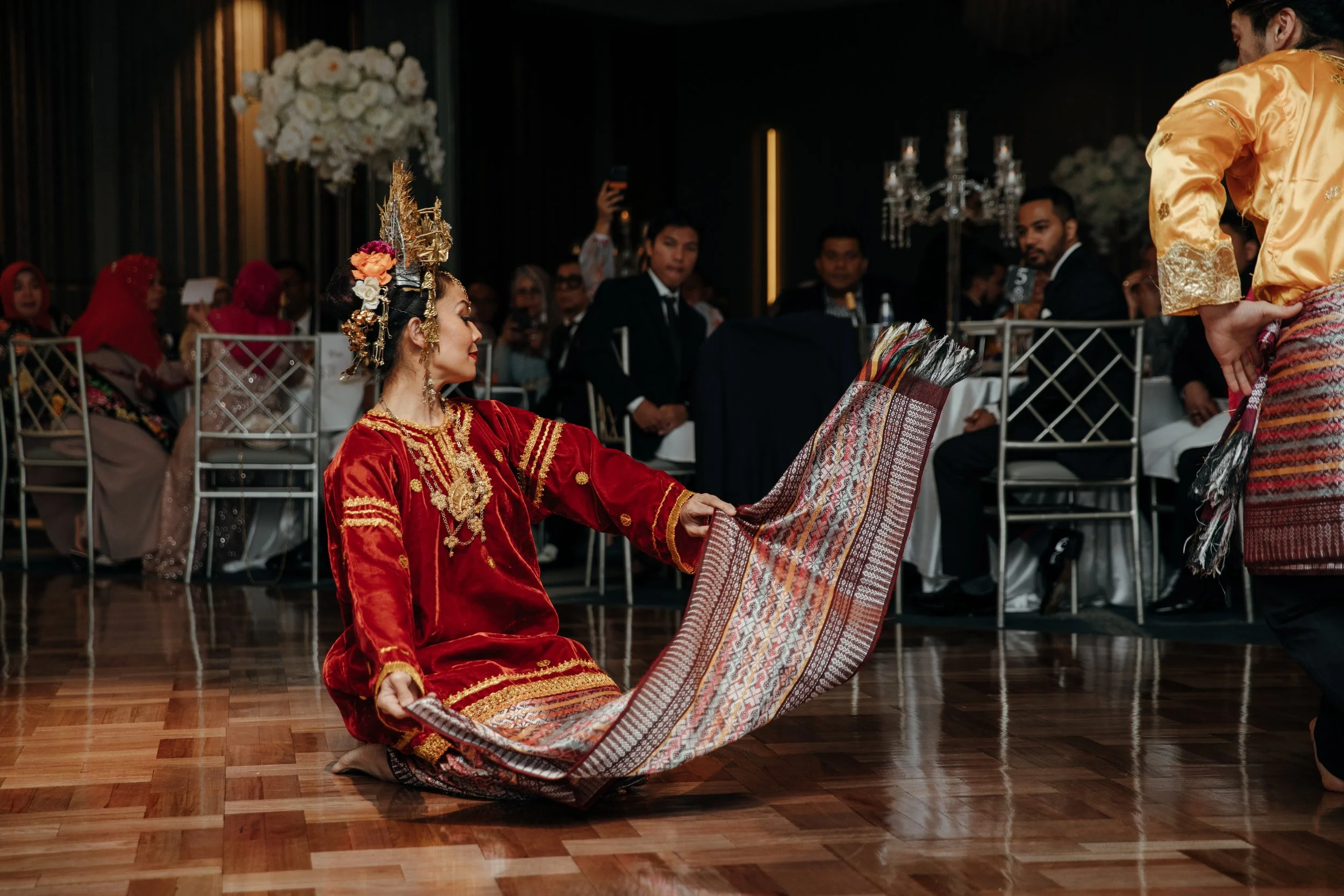A woman dressed in traditional Indonesian attire performs a dance on a wooden floor at a formal event, with seated guests watching in the background.