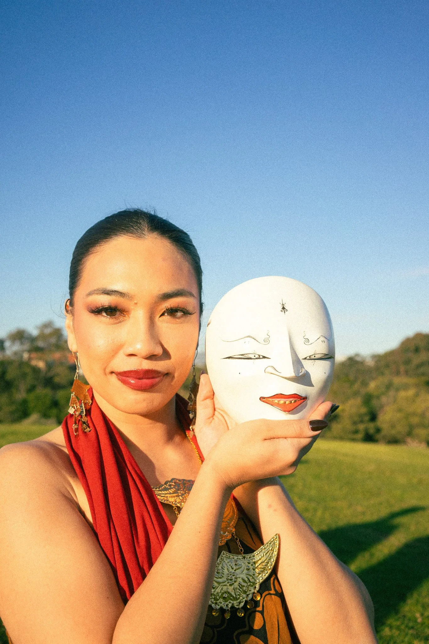 A woman in traditional Indonesian attire holding a decorative mask with a smiling face, outdoors during the daytime with a clear blue sky and greenery in the background.