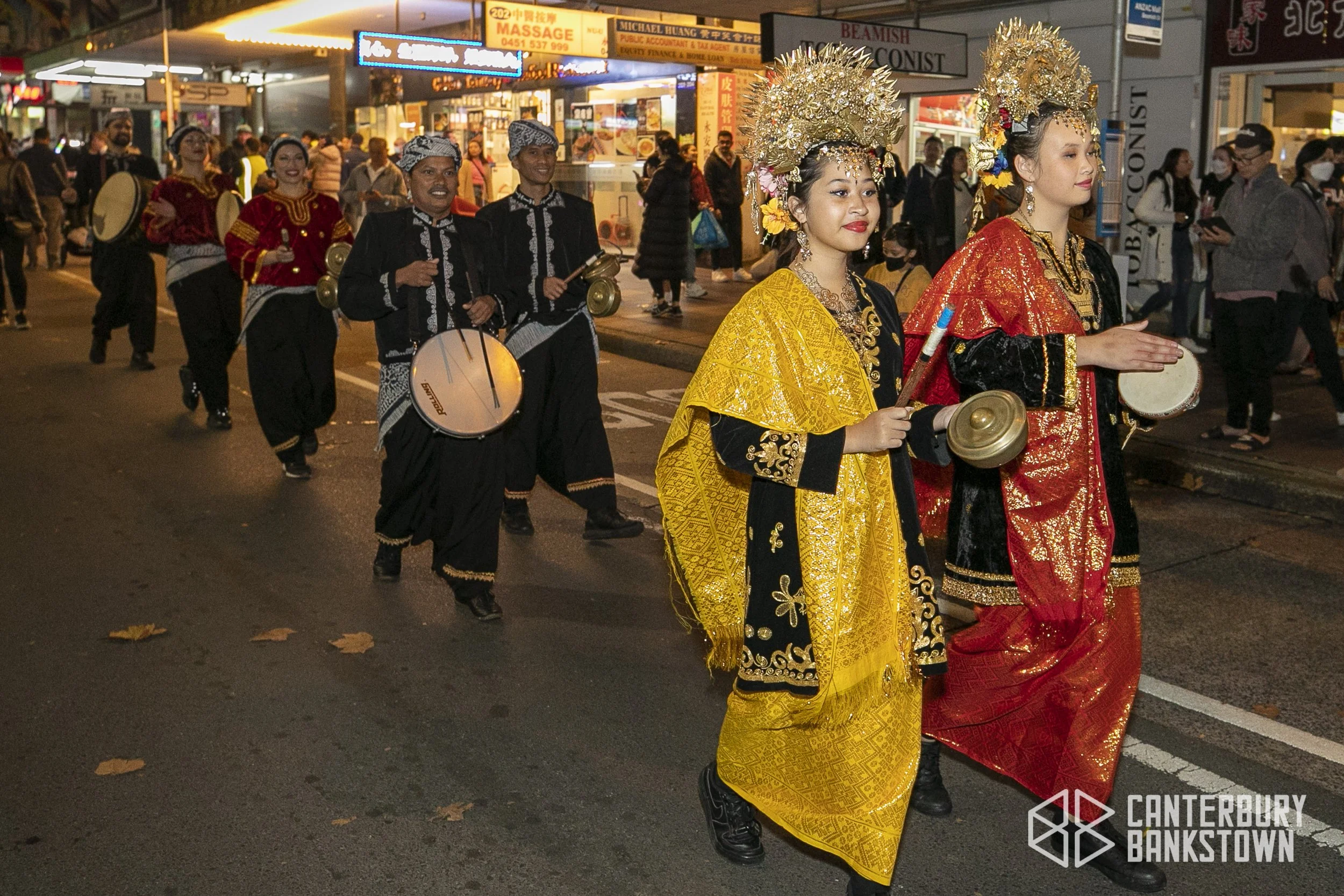A group of people dressed in traditional costumes, including two women in ornate gold and red outfits with headpieces, leading a parade on a city street at night. Behind them, men wearing black outfits with white patterns are playing drums and cymbals, performing as part of the procession. The street is busy with spectators and shops illuminated with neon signs.